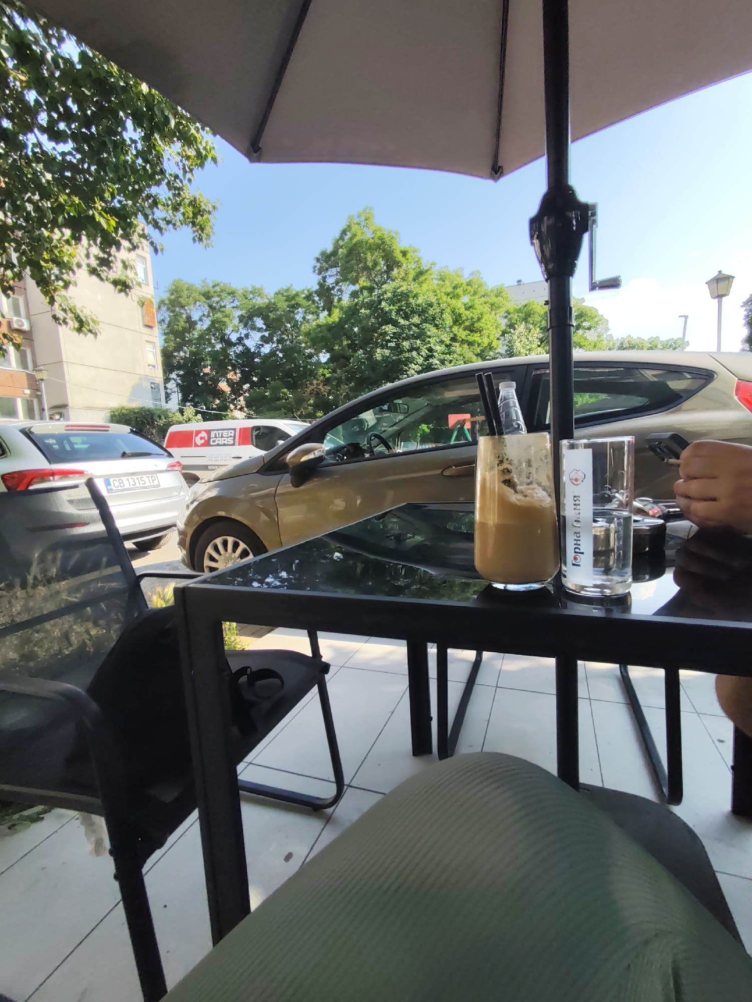 The image captures an outdoor cafe scene on a clear, sunny day in the city of Plovdiv, Bulgaria. The perspective is from a seated individual whose lower body, clad in light green/grey ribbed trousers, is visible in the lower right foreground. A black rectangular table stands directly in front, bearing two glasses. The left glass, tall and half-full, contains a frothy, light brown beverage with two black straws and a small plastic bottle inserted. The right glass holds clear water and features a white label with a red circular logo and the Cyrillic text "ГОРНА БАНЯ" (GORNA BANYA). A person's right hand, holding a dark object, is visible to the right of the water glass.
In the midground, a bronze/gold compact car is parked facing right, partially obscured by the table and a central black metal umbrella pole. To its left, a silver Skoda Octavia is parked, displaying the Bulgarian license plate "CB 1315 TP." Behind these vehicles, a white van with a prominent red "INTER CARS" logo is visible. The light-colored canopy of a large outdoor umbrella shades the scene from above.
The background consists of multi-story buildings with windows and balconies, interspersed with lush green trees and foliage under a clear blue sky. An exterior air conditioning unit is visible on one building. A streetlamp is partially visible on the far right. The ground beneath the table and chairs is tiled with light-colored square pavers.