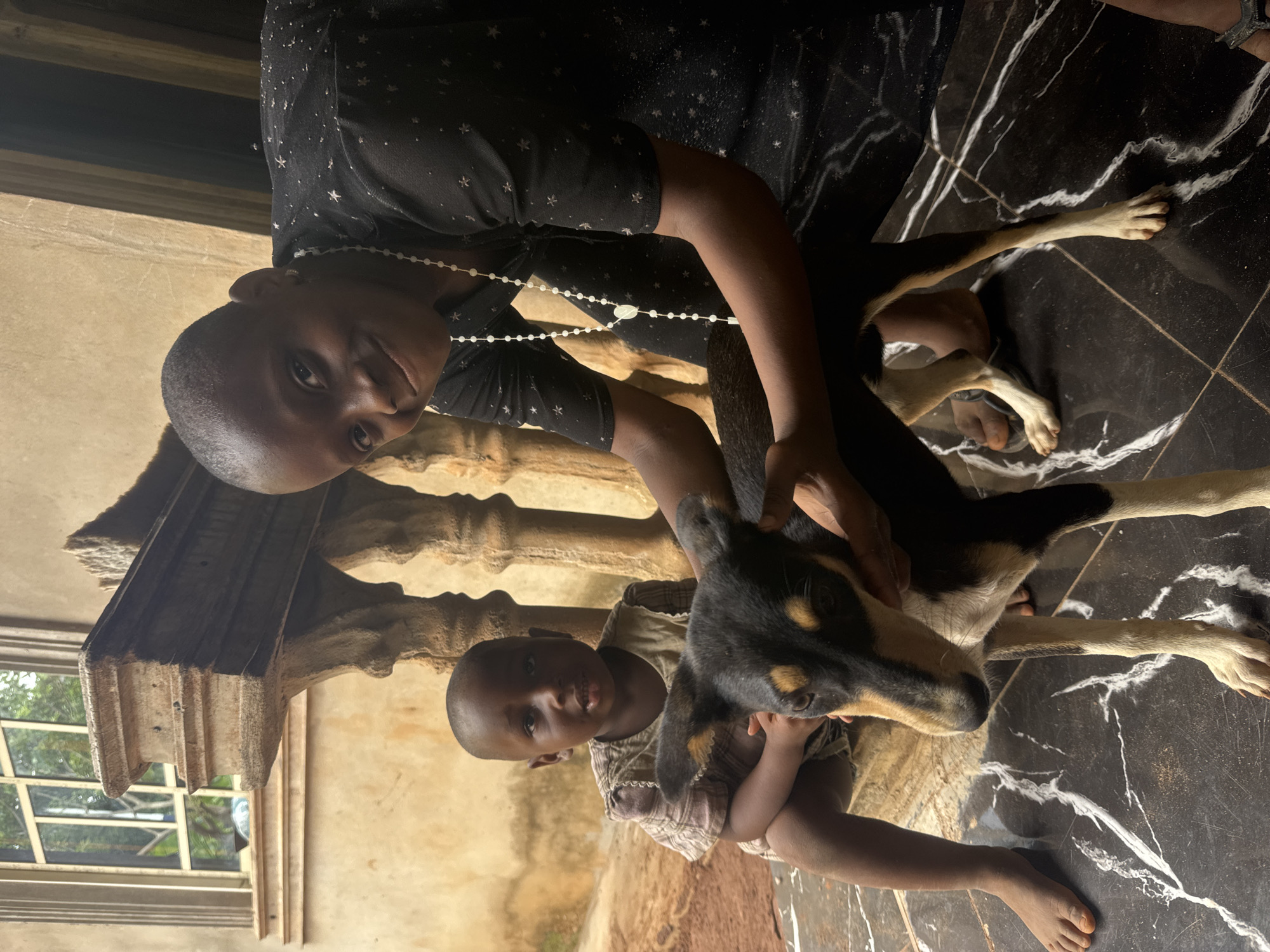 This image, taken in Eziudo, Nigeria, features two children and a dog interacting indoors. On the left, an older child with dark skin and short hair wears a black long-sleeved top adorned with white star patterns and a white beaded necklace. This child is looking directly at the camera with a neutral expression, with one hand gently resting on the dog's back.
Adjacent to the older child and slightly forward, a younger child, also with dark skin and short hair, is seated on the floor. This child wears a light-colored, possibly plaid or striped, short-sleeved top and looks towards the camera. Their hands are positioned on the dog's head and neck area.
Between the two children is a medium-sized dog with a black, tan, and white coat. The dog's head, primarily black with tan markings around the eyes and snout, is visible, and it appears to be looking towards the camera or slightly to the side, being petted by both children.
The setting features a distinctive black floor with prominent white marbled or veined patterns. In the background, an ornate, light-colored architectural pillar or column is visible, along with a window through which green foliage can be seen, indicating it is daytime. The overall scene depicts a gentle and engaged interaction between the children and their dog. There is no visible text in the image.