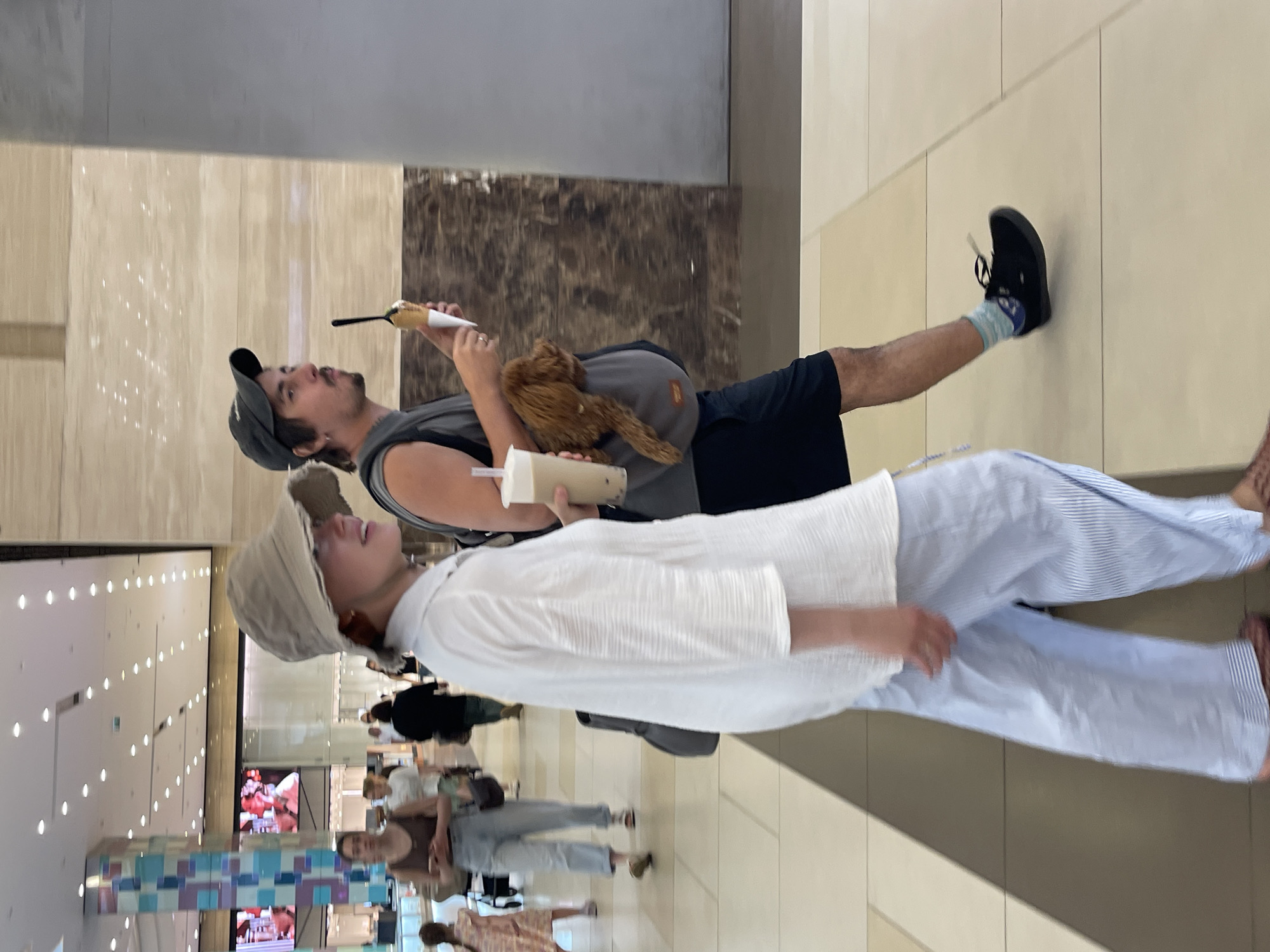 The image captures a moment in a brightly lit, indoor shopping mall with a polished floor and contemporary architecture, including tall columns and storefronts in the background. In the foreground, a couple is walking side-by-side. The woman, on the left, wears a light-colored, wide-brimmed hat, a loose white collared shirt, and light blue striped pants. She is holding a clear plastic cup with a beverage. The man, on the right, wears a dark baseball cap, a dark sleeveless shirt, and dark shorts. He is carrying a small, light brown dog in a grey sling carrier worn across his chest and is holding an ice cream cone. They appear to be casually strolling through the mall. Other shoppers are visible in the background, some looking at displays or walking in different directions. The overall impression is a typical scene within a modern retail environment.