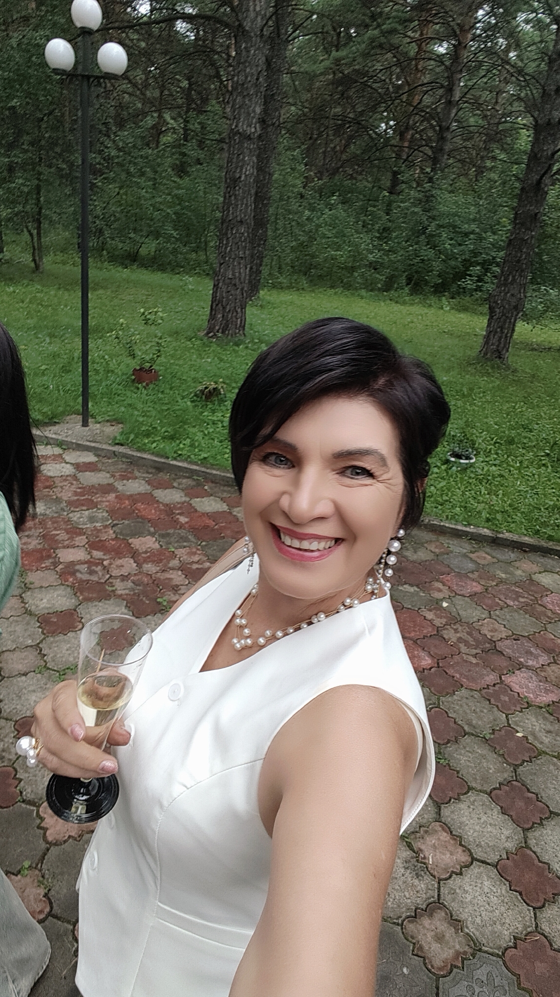 A woman, appearing to be in her late 50s or early 60s, with short, dark hair and blue eyes, is taking a selfie outdoors. She is smiling and holding a champagne flute in her left hand. She is wearing a sleeveless white top, a pearl necklace, and large pearl earrings. The background suggests a park-like setting with trees and grass, and a paved walkway with reddish-brown and gray interlocking stones. A lamppost with three white globes is visible to her left, and another person's dark hair is partially visible in the lower left corner of the frame. The lighting suggests it is daytime, possibly overcast. The location is Beryozovka, Russia.