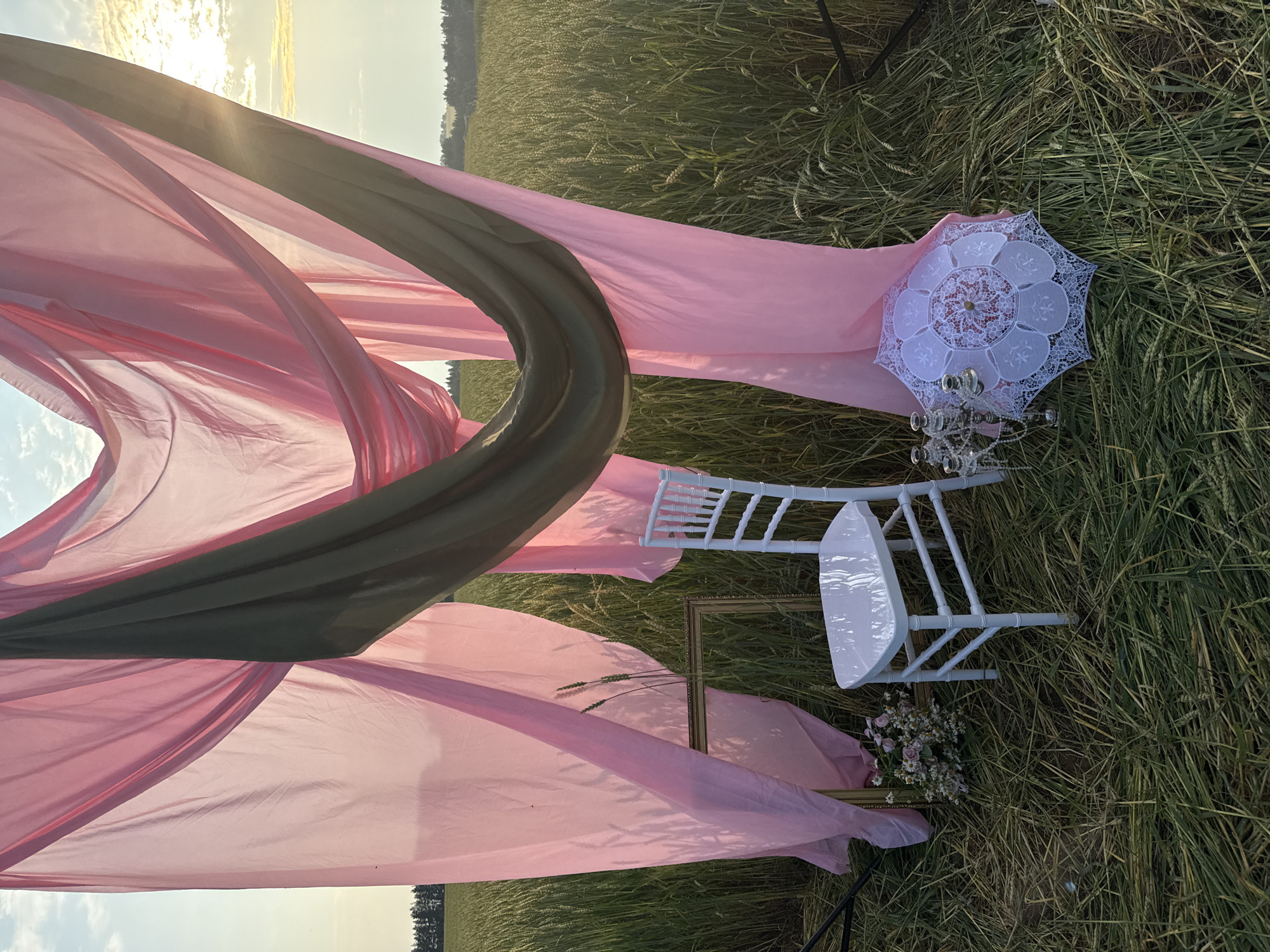 An outdoor, low-angle, eye-level, medium full shot shows a white chair placed in a field of dried grass and wheat. Pink and olive green sheer fabric are draped across the top of the frame, obscuring the sky. To the left of the chair is a golden rectangular frame and a small bouquet of flowers. To the right of the chair is a silver candelabra and a white lace parasol. The image appears to be taken at sunset, with the sun casting a warm glow on the scene. The location is in the city of Foki, Russia. There are no people in the image.