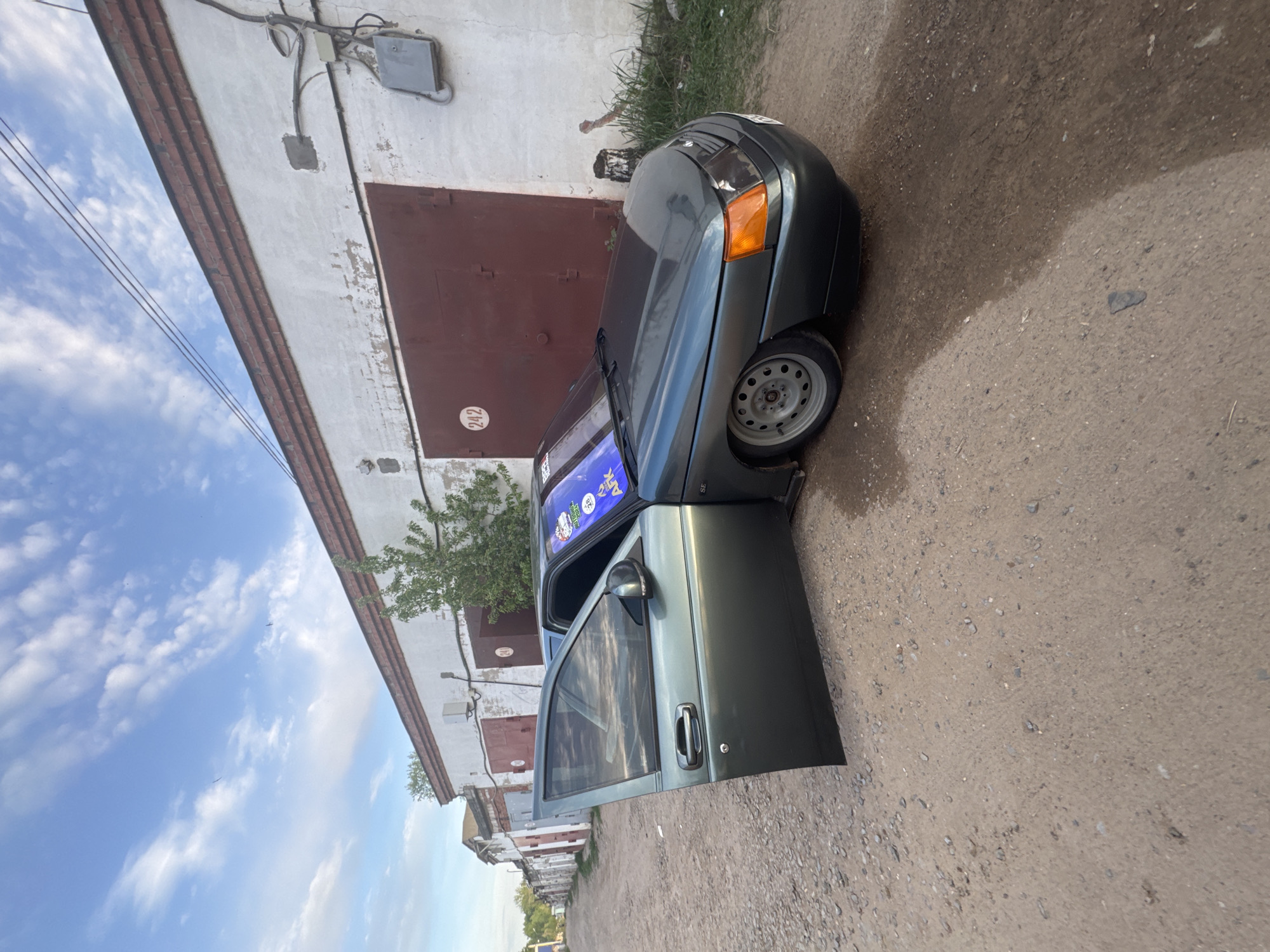 The image depicts a green car parked on a gravel surface, with the driver's side door open. The car is positioned in front of a white building with a brown garage door, the garage door has the number 242 on it. Overhead electrical lines and a utility box are visible on the wall of the building. The sky is blue with scattered clouds. The location is likely Chaikovsky, Russia.