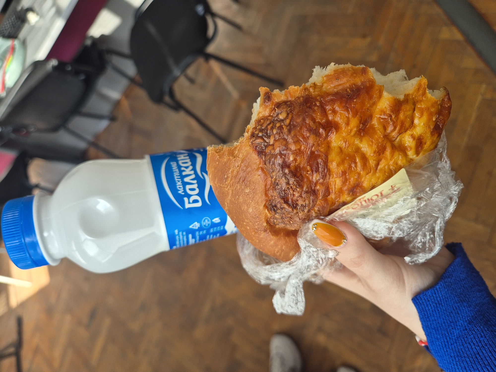 A hand holds a partially eaten piece of cheesy bread wrapped in plastic, juxtaposed against a bottle of Balkan dairy drink. The background shows a wooden floor, black office chairs, and a partial view of a table. This likely represents a quick meal setting, possibly in a cafe or office, but the specific location in Pavlikeni, Bulgaria cannot be determined from the image alone.
