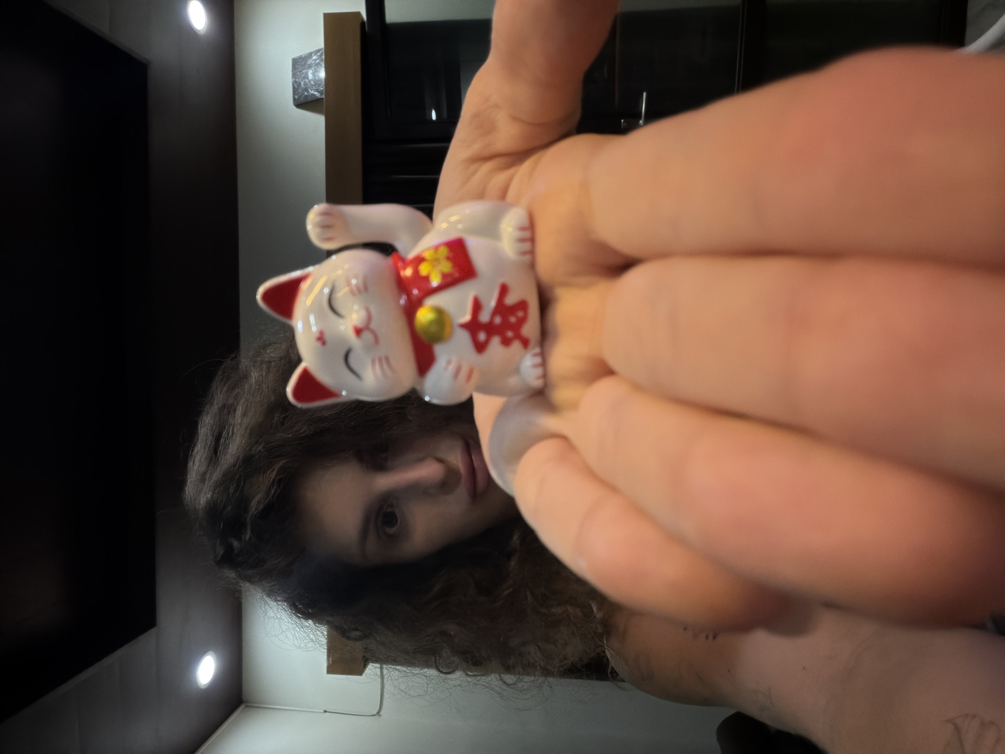 The image is a close-up, vertically oriented shot, rotated 90 degrees counter-clockwise. A person with long, dark, curly hair and a neutral expression is visible on the left side of the frame, looking towards the camera. Their right hand and fingers are prominent, holding a small white Maneki-neko (lucky cat) figurine centrally.
The Maneki-neko has red ears and is adorned with a red collar, a golden bell, and a red bib featuring a golden Chinese character, "福" (fú), which translates to "fortune" or "good luck." The figurine has a peaceful, smiling face with closed eyes, and its right paw is raised. The person's fingers and palm appear large in the foreground, framing the figurine.
The background reveals a dimly lit indoor setting, with dark walls and a ceiling. Two bright, circular, wall-mounted light fixtures are visible, providing illumination to parts of the room. Dark rectangular shapes, possibly furniture or architectural elements, are subtly present in the background. The overall impression is a casual, intimate shot, likely a selfie, focusing on the person presenting the lucky cat. No specific time of day or weather is discernible from the indoor scene.