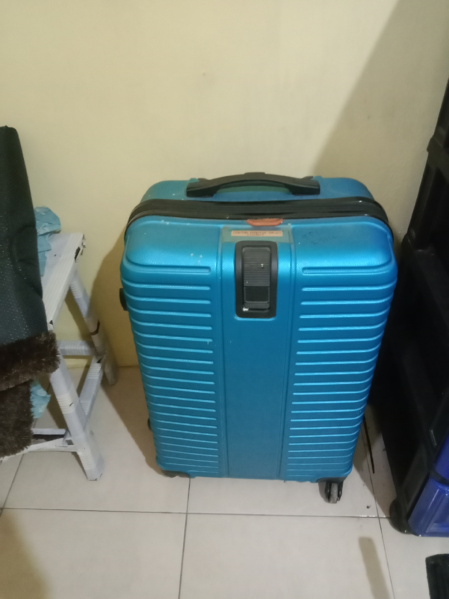 A bright blue, hard-shell suitcase with black trim and wheels is positioned in the center of the frame. It has a retractable handle on top and a black, recessed latch mechanism with an orange accent on the front. To the left of the suitcase, a portion of a dark-colored garment with a furry trim is visible, draped over a white, folding stool. The stool has a triangular structure. The background consists of a light-colored wall. The floor is tiled in a light beige color with dark grout lines. The lighting suggests an indoor environment, possibly a room in Kutosari, Indonesia, with a shadow cast by an unseen object on the right side of the suitcase and wall.