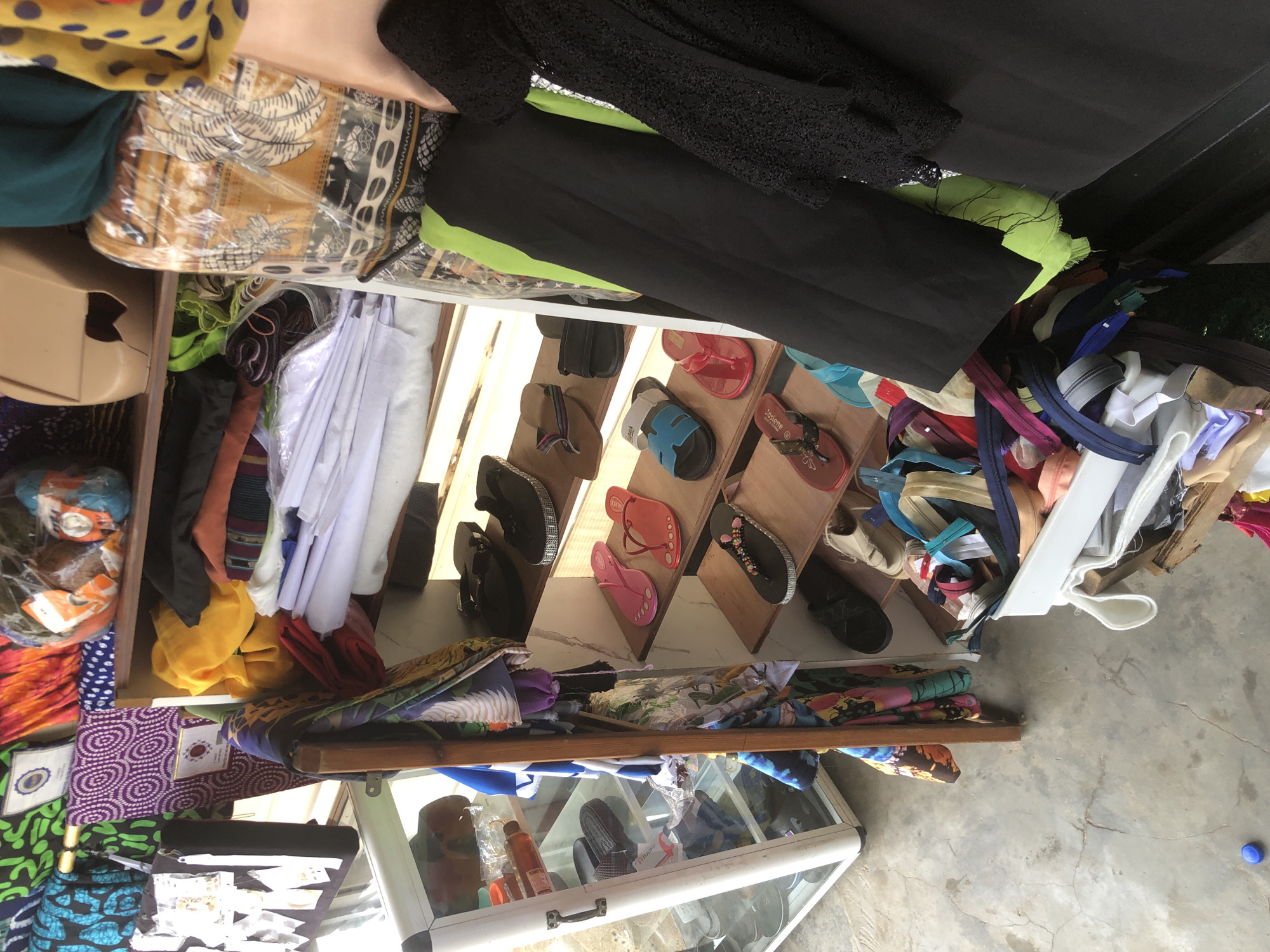 This image captures a vibrant and densely packed market stall or small shop, likely in an outdoor or semi-outdoor setting in Ado, Nigeria. The scene is characterized by a wide array of merchandise, primarily textiles, footwear, and sewing supplies, displayed for sale.
**Subjects:**
The primary subjects are numerous objects arranged on various shelves and surfaces. Dominant among these are colorful fabrics, including patterned African prints (Ankara-style), solid colored cloths in yellow, green, black, and white, and what appears to be black lace fabric. Many of the fabrics are neatly folded or rolled, some packaged in clear plastic.
Footwear, specifically sandals and flip-flops, are prominently displayed on a multi-tiered wooden shelf unit. These shoes come in various colors such as red, pink, blue, black, and beige, with some featuring decorative embellishments. Below these, a large collection of zippers in assorted colors (blue, pink, beige, black, white, purple) is visible, likely in a bin or on a lower shelf.
A glass display case at the bottom contains additional items, including bottles, one of which appears to be a "Honey Glow" brand product, and more footwear. Other visible objects include small white tags/labels on fabrics, a beige plastic container, and a small blue bottle cap on the floor.
**Activity or Event:**
The image depicts a retail setting, suggesting the ongoing activity of commerce and trade. Goods are presented for customers to browse and purchase.
**Notable Details:**
The bright, natural lighting indicates it is daytime. The floor is concrete with visible cracks, typical of market or open-shop environments. Despite the large quantity of items, there's an attempt at organization, with shoes displayed on dedicated risers and fabrics folded. The overall impression is one of abundance and variety.
**Visible Text:**
"Honey Glow" is faintly visible on a bottle within the glass display case. "J'adore" or "S'adore" is visible on the strap of a pink flip-flop on the wooden display. Smaller, unreadable text is present on various labels and tags throughout the stall.