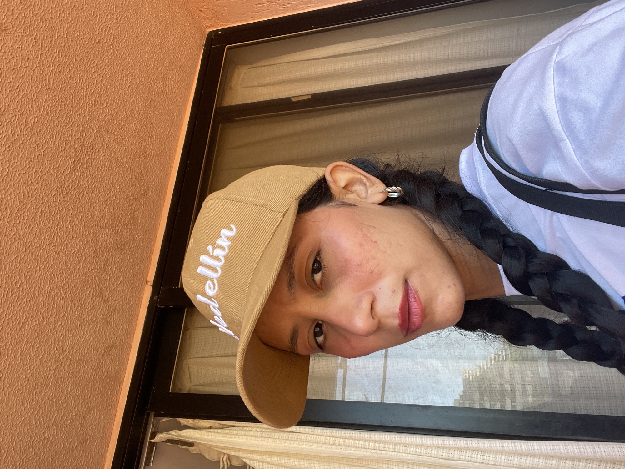 A woman with dark braided hair and a tan baseball cap with white lettering that reads "Medellin" poses for a selfie outdoors. She is wearing a white shirt and silver hoop earrings. The background consists of a window with sheer curtains and an orange textured wall. The scene is likely a residential exterior in Porlamar, Venezuela, possibly a balcony or patio area.