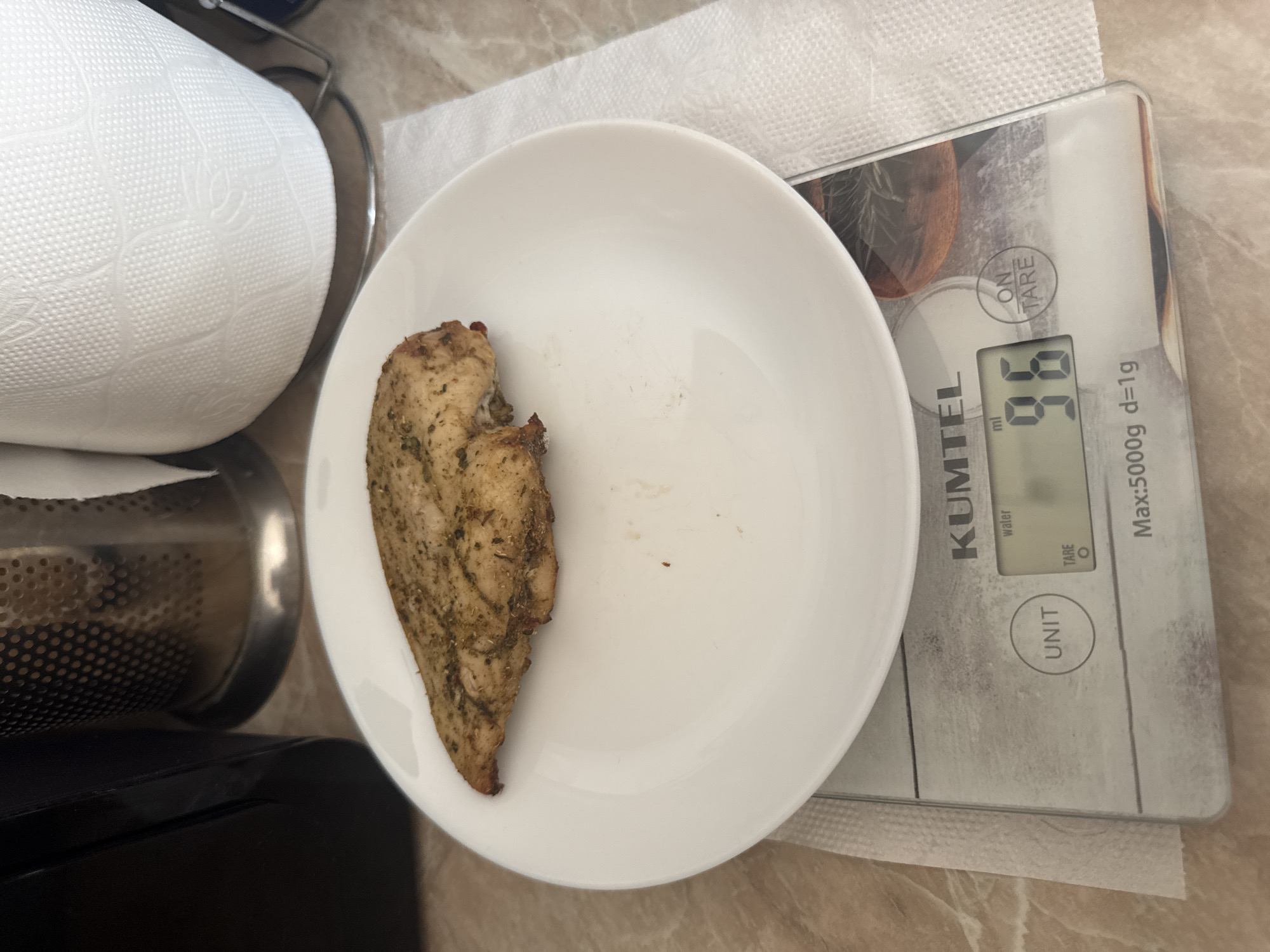 An overhead, close-up shot captures a piece of seasoned chicken breast resting on a white plate, which in turn sits on a digital kitchen scale. The scale displays a weight of "96" grams, with "ml" and "water" visible on the screen. The background shows a countertop, a roll of paper towels, and a metallic object that appears to be a utensil holder. The scene is likely set in a kitchen environment in Tbilisi, Georgia.