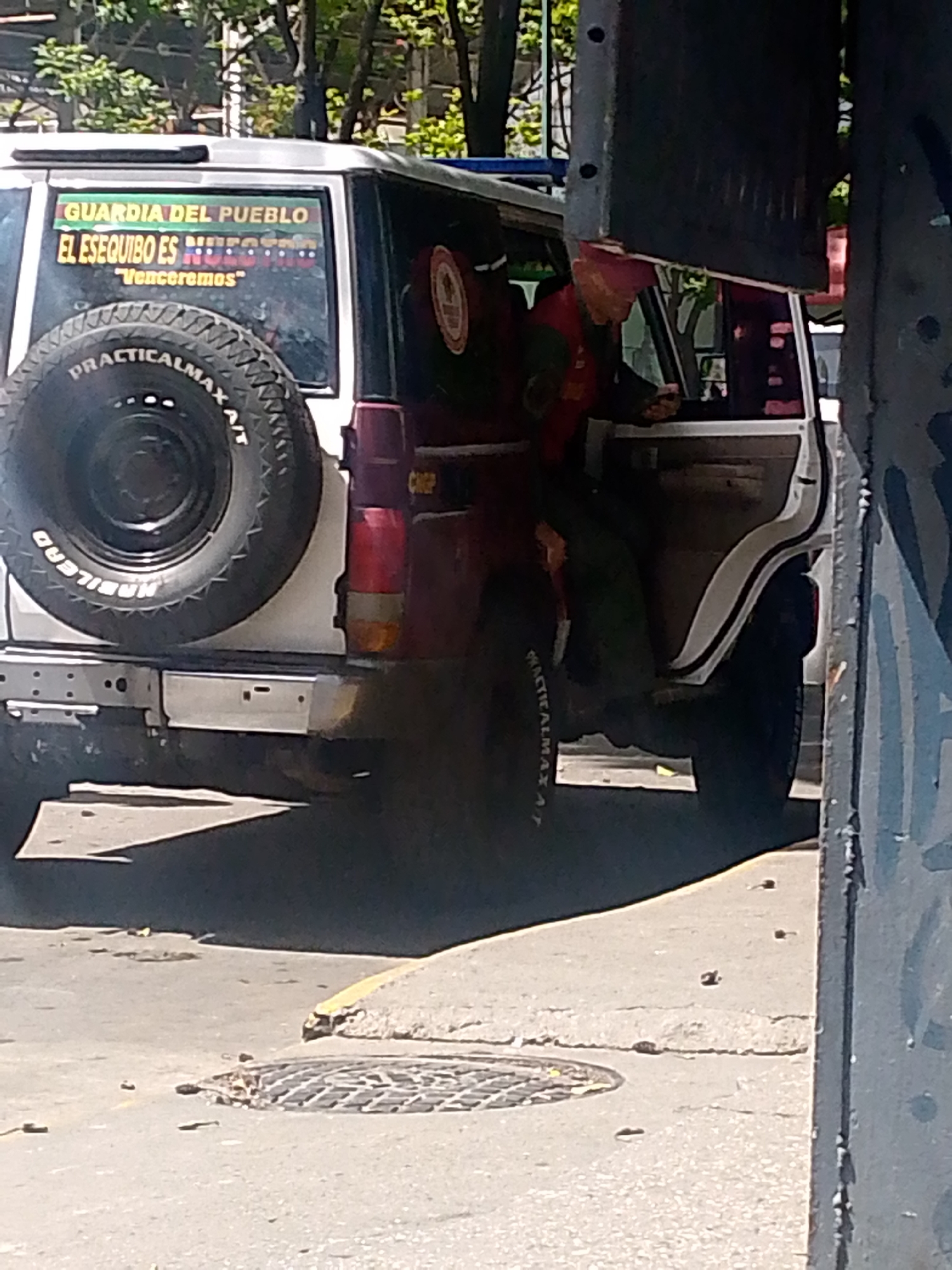 A white and maroon jeep with the words "GUARDIA DEL PUEBLO" and "EL ESEQUIBO ES NUESTRO" with the phrase "Venceremos" written on its side. The vehicle is parked on a city street in Caracas, Venezuela. There is a man inside the vehicle, wearing a uniform. A metal grate is visible on the street near the vehicle. A metal pole, possibly a streetlight, obscures part of the view.