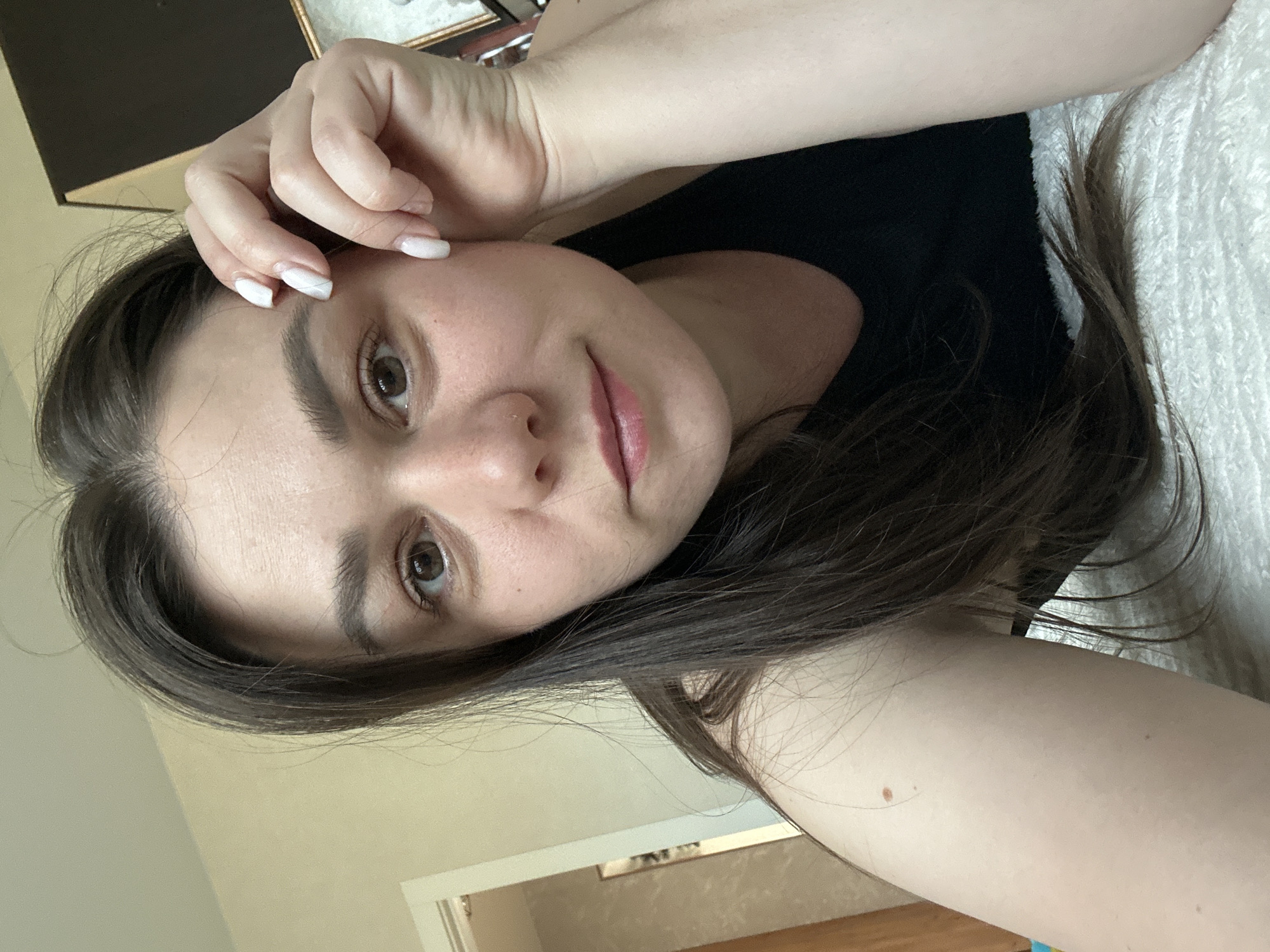 A woman is lying down and has her right hand touching her forehead. She has dark hair and brown eyes. She is wearing a black top. Her fingernails are painted white with a swirl design. She has a nose piercing. She is on a white, fluffy surface. There is a door and window in the background, and furniture. It is not possible to determine if the photo was taken in the city of Chaikovsky, Russia.