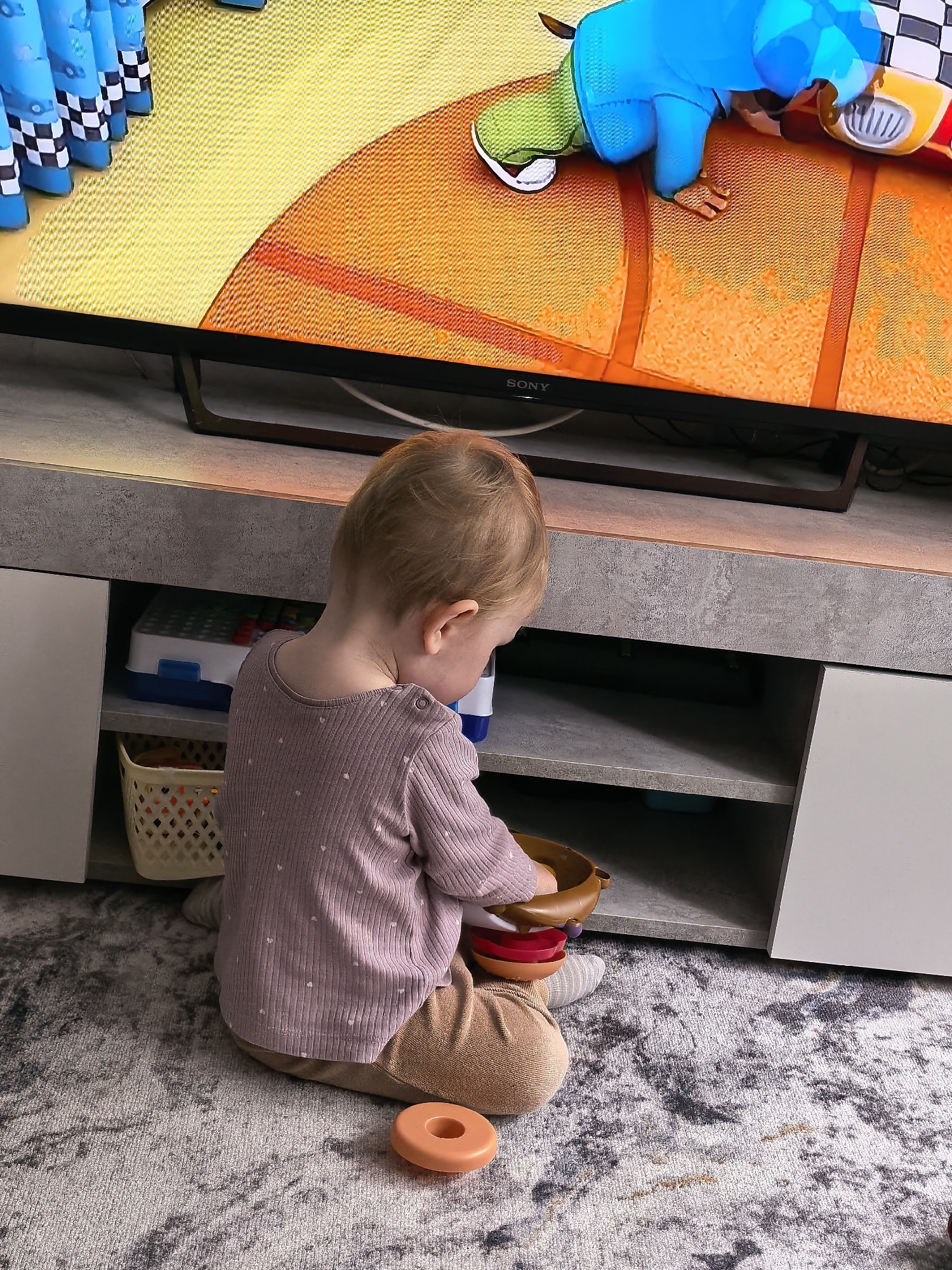 An indoor scene depicts a young child seated on a grey, white, and black patterned rug in front of a television and a grey media console. The child, viewed from behind, has light-colored hair and is wearing a long-sleeved light purple top with small white polka dots and light brown pants. The child is actively engaged with several toys, holding a brown bowl-shaped object and appearing to manipulate smaller, colorful bowls (red, possibly others) within or near it. An orange ring-shaped toy rests on the rug to the child's right.
A SONY brand flat-screen television prominently displays an animated scene featuring brightly colored characters or objects on a textured orange and yellow background. On the left side of the screen, multiple blue figures with black and white checkered bases are aligned. On the right, a character in a blue top and green bottom is positioned on the orange surface next to a red and yellow object resembling a helmet or vehicle part.
The media console, made of a textured grey material, features open shelving compartments. The upper left shelf contains a white and blue container, possibly holding building blocks. A light-colored woven basket is visible on the lower left shelf. The shelves on the right side appear largely empty. Cables are visible behind the television and within the console's rear section.
There are no discernible visual elements within the image to indicate its specific geographical location, thus the city of Sterlitamak, Russia, cannot be confirmed.