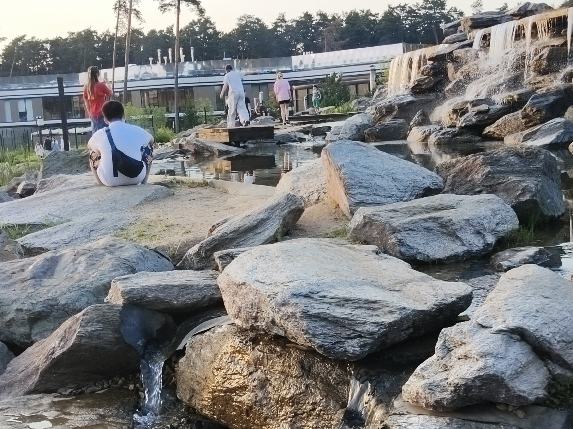 The image captures an outdoor recreational area in the city of Unknown, Unknown, under late afternoon or early evening lighting. The scene features a constructed landscape of numerous large, grey, angular rocks and water elements. On the right, a multi-tiered artificial waterfall cascades over a formation of rocks into a series of pools and channels. Smaller streams of water flow between rocks in the foreground and mid-ground.
Several individuals are present. In the left foreground, a person wearing a white t-shirt and dark patterned shorts, with a black sling bag across their back, sits on a large flat rock, facing away from the camera. Immediately behind this individual, another person with long, light brown hair, dressed in a red top and light-colored pants, stands holding a smartphone raised as if photographing or videoing the background. Further back, centrally, a person in light-colored long pants and a light top walks across a wooden structure spanning a water channel. To their right, another person in a pink t-shirt and dark shorts walks along a stone path or steps. A younger individual in a green t-shirt and light shorts is visible further to the right on this path. In the background, a large, modern building with a light-colored facade and extensive window panels spans horizontally, backed by a dense line of trees. The lighting casts warm, golden reflections on the water and wet rock surfaces.