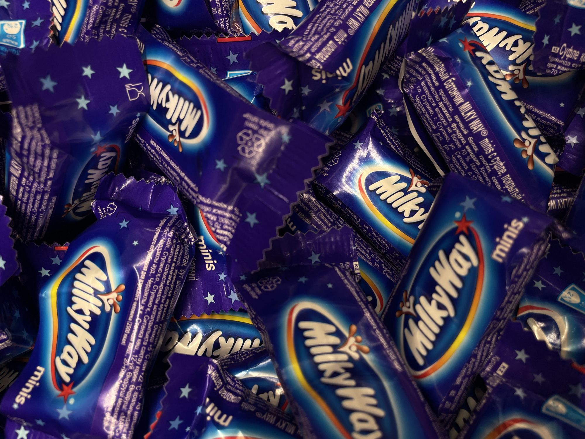 The image is a close-up, overhead shot of a pile of individually wrapped Milky Way candy bars, specifically the "minis" size. The wrappers are predominantly deep blue with the prominent "Milky Way" logo in white, outlined in red and yellow, featuring the characteristic splash of chocolate. Scattered across the wrappers are small, light blue stars. The lighting is bright and somewhat harsh, creating reflections on the plastic packaging. The candies are densely packed, creating a textured, almost abstract composition of blues, whites, reds, and yellows. There is no visible indication of a specific location in Rostov-on-Don, Russia, nor are there any people or specific environmental elements like buildings or streets. The focus is entirely on the product packaging itself.