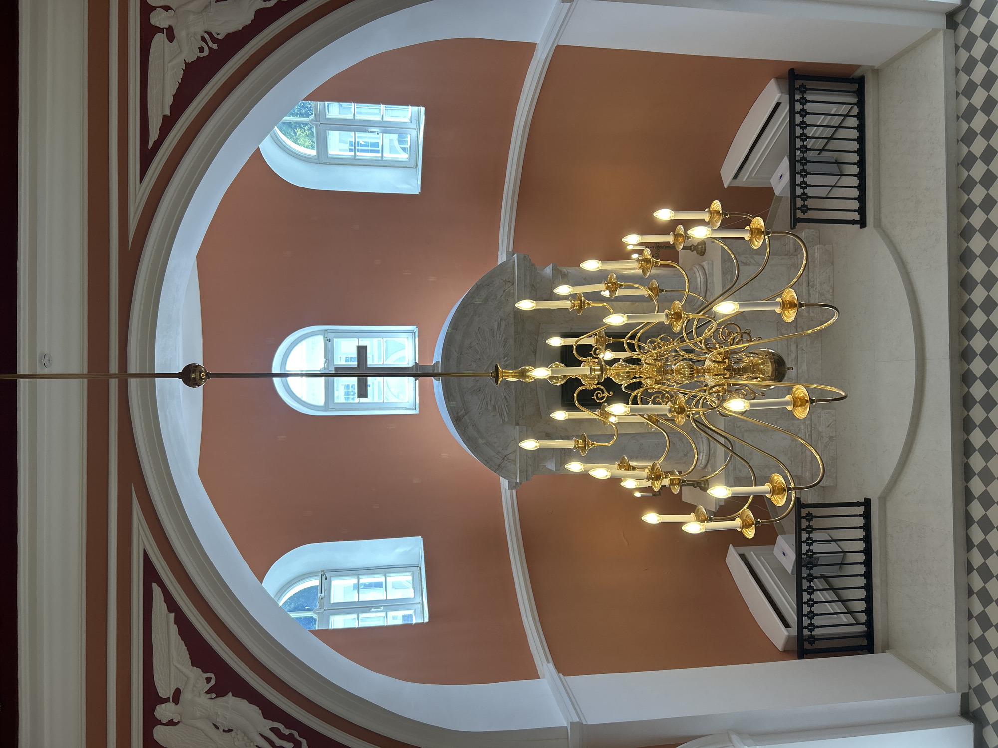 This image depicts the interior of what appears to be a church or a grand building, possibly located in the village of Dobasnen, Dobasnen Rural Council, Belarus. The central focus is a large, ornate, gold-colored chandelier with numerous candle-like lights, suspended from the ceiling. It hangs in front of an arched alcove containing a cross visible through an arched window.
The walls are painted a warm, peachy-orange color, and the ceiling features decorative architectural elements, including white molding and relief carvings of angelic figures on a deep red background. Three arched windows with white frames are visible, two on either side of the central alcove and one slightly above and to the left. The windows appear to have a grid pattern and let in natural light.
Below the chandelier, a white marble structure serves as a backdrop. In front of this structure, to the left and right, are black metal railings. Behind the railings, on the floor, are two white air conditioning units or heaters. The floor area in the foreground has a checkered pattern of grey and white tiles. The overall impression is one of architectural grandeur and religious or historical significance. There are no people visible in the image.
