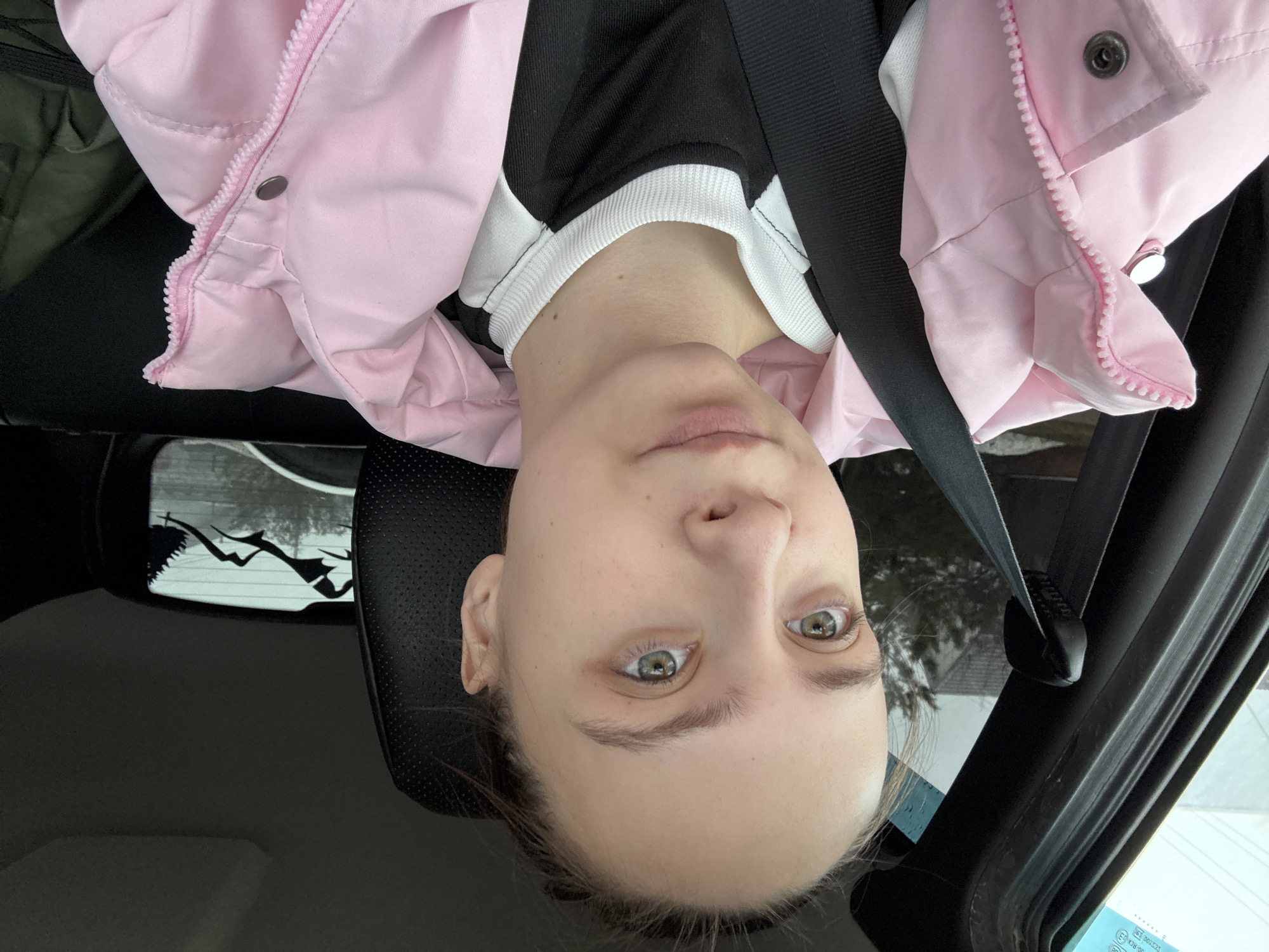 A person wearing a pink jacket and a black and white top is in a car, secured by a seatbelt. The location appears to be Прикамский, Russia, based on the information available. The person is looking directly at the camera. A mountain scene decal can be seen on the rear window of the car.