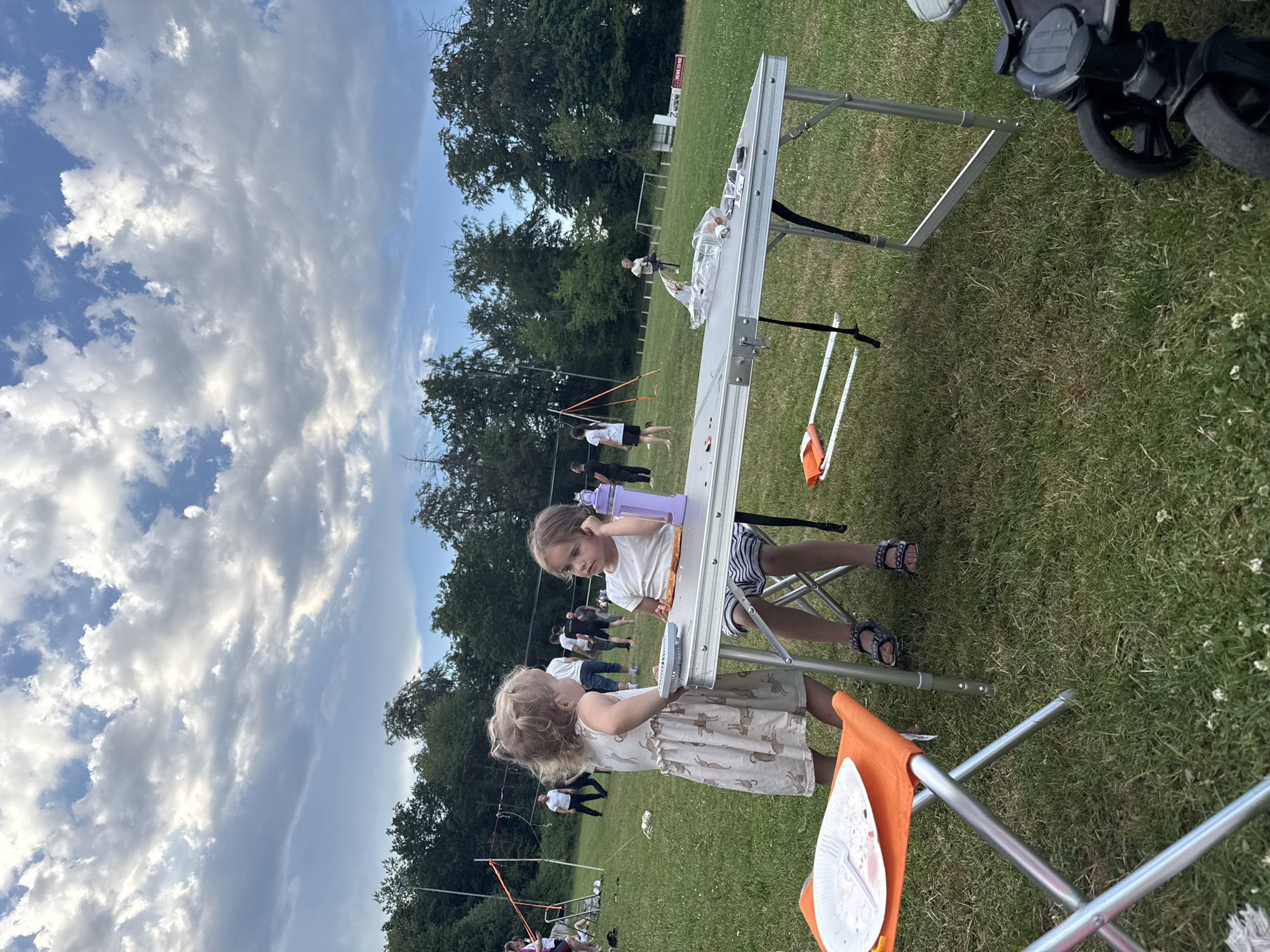The image depicts an outdoor scene in Lumda, Germany, likely a park or sports field, on a cloudy day. In the foreground, two young girls are interacting near a metal folding table. The girl on the left, wearing a light-colored dress with a pattern, is standing and facing the girl on the right. The girl on the right, wearing a white t-shirt and striped shorts, is seated and appears to be looking at something on the table, which holds a slice of pizza and a purple bottle. An orange folding stool with a plate of food is positioned to the left of the table.
In the background, several adults and children are visible on a grassy field. Some appear to be engaged in activities related to volleyball, with a net visible in the mid-ground. Trees and foliage form a backdrop along the edge of the field. Further in the distance, structures like goalposts and a white building are present, suggesting a sports or recreational facility. The overall impression is of a casual outdoor gathering.
