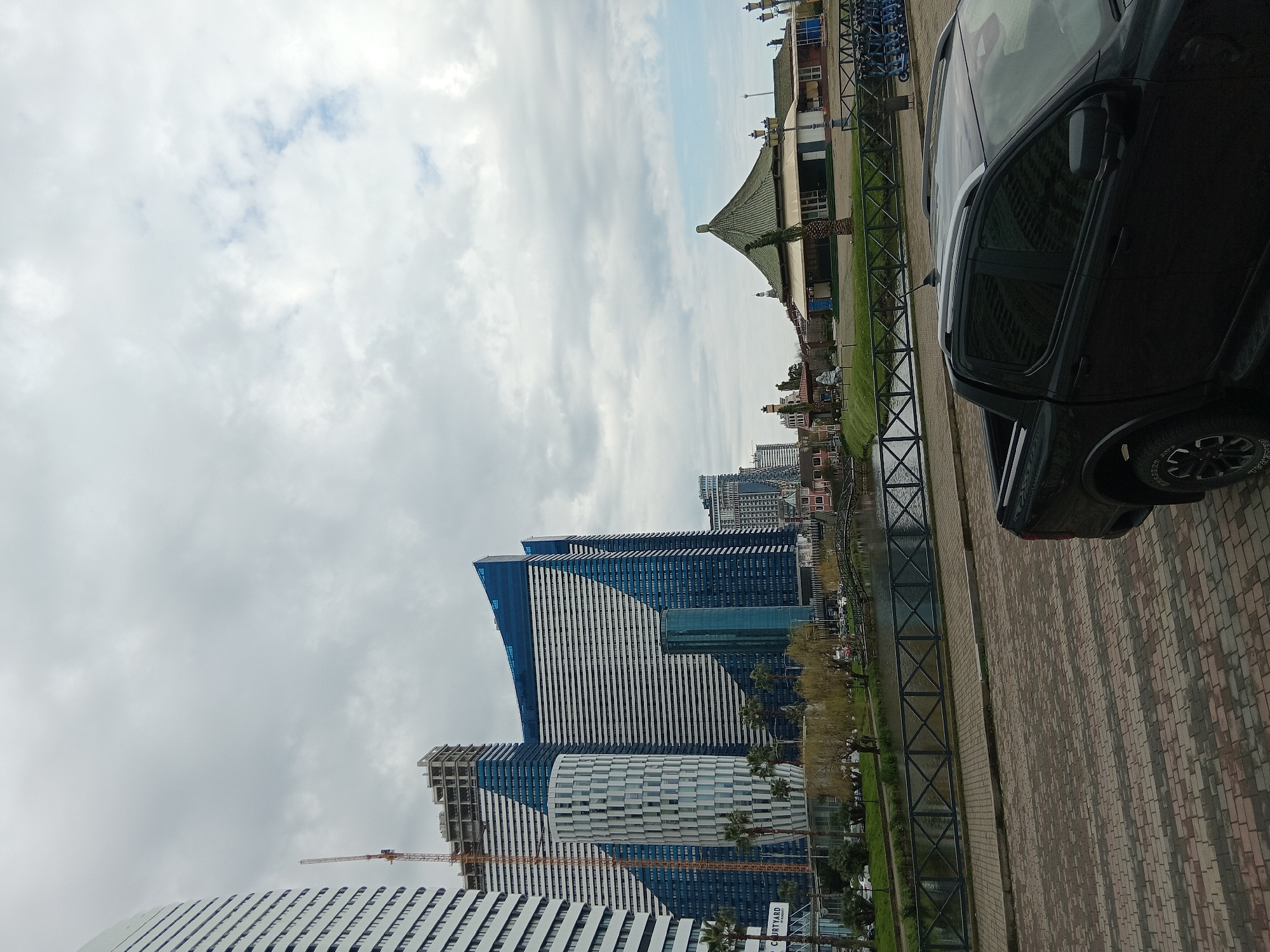 The image depicts a street scene in Batumi, Georgia, captured on an overcast day. Modern high-rise buildings with distinctive blue and white patterned facades dominate the view, with construction cranes visible amidst the architectural structures. A black SUV occupies a portion of the right side of the frame, parked along a brick-paved street adjacent to a metal railing that separates the walkway from a narrow strip of green space alongside a body of water. Further in the background, traditional buildings with peaked roofs and other urban features are visible, set against a cloudy sky. The scene shows a mix of contemporary architecture and urban infrastructure in Batumi.