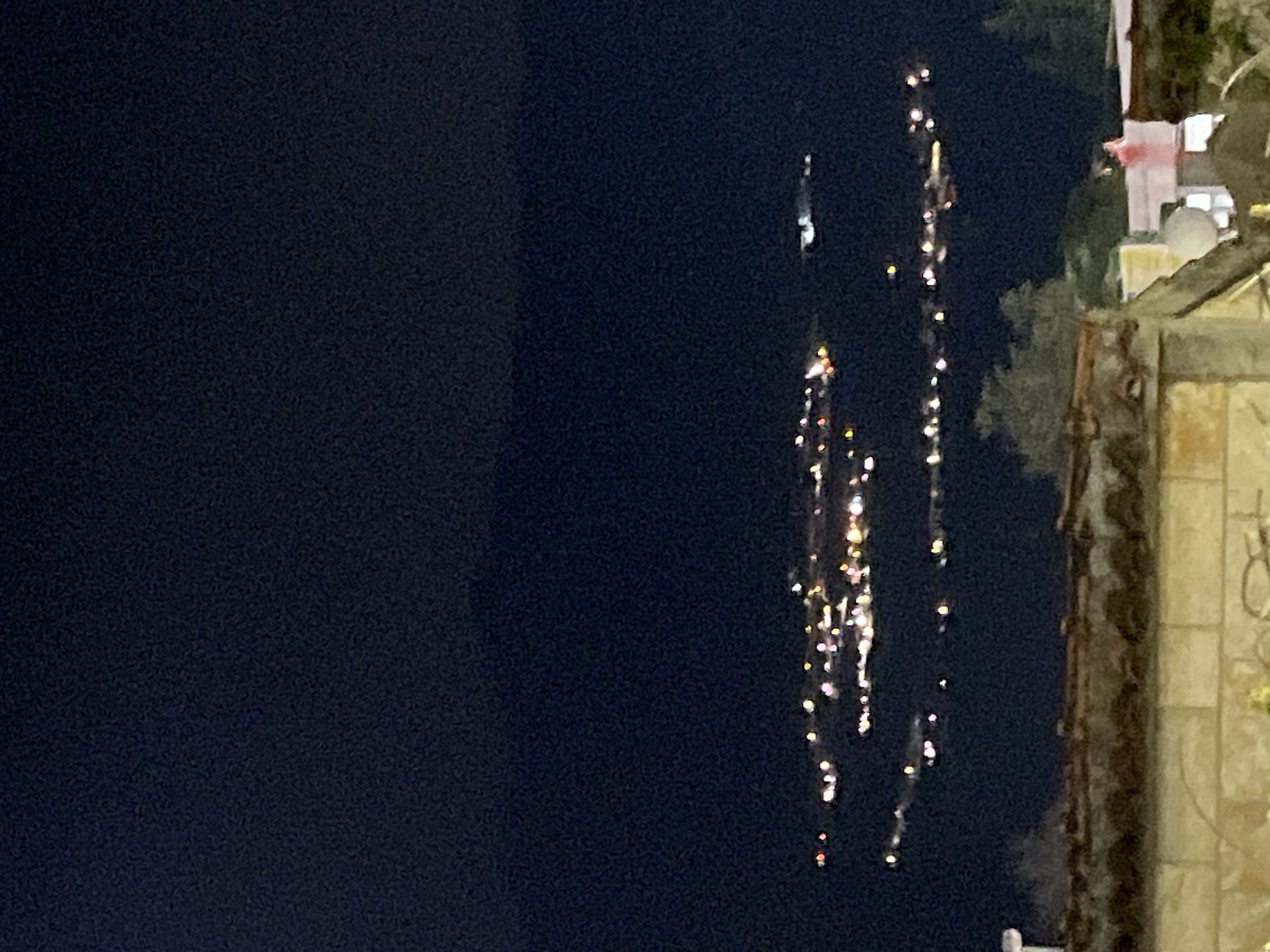 The image depicts a nighttime scene in Budva, Montenegro, as evidenced by the presence of buildings and urban lighting. Two vertical columns of bright lights, presumably from decorative displays or streetlights, stand out against a dark background. The right side of the image includes the partial view of a building or structure. The sky is dark, suggesting it is after sunset. The overall impression is a view of an urban area at night.