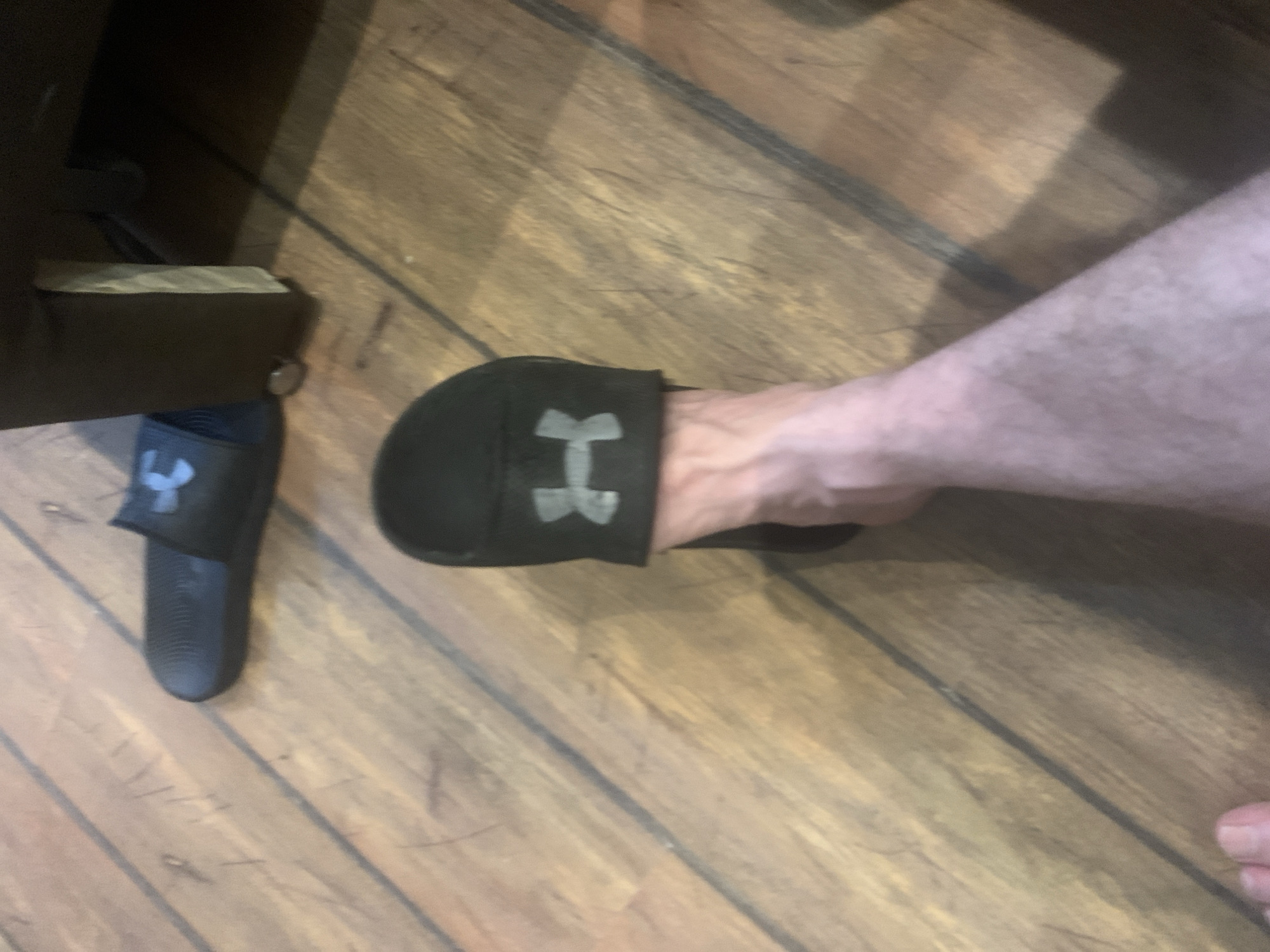 A person's feet are visible on a wooden floor. One foot is wearing a black Under Armour slide sandal. Another similar sandal is visible in the background. A dark brown piece of furniture is to the right of the image. The scene appears to be indoors.