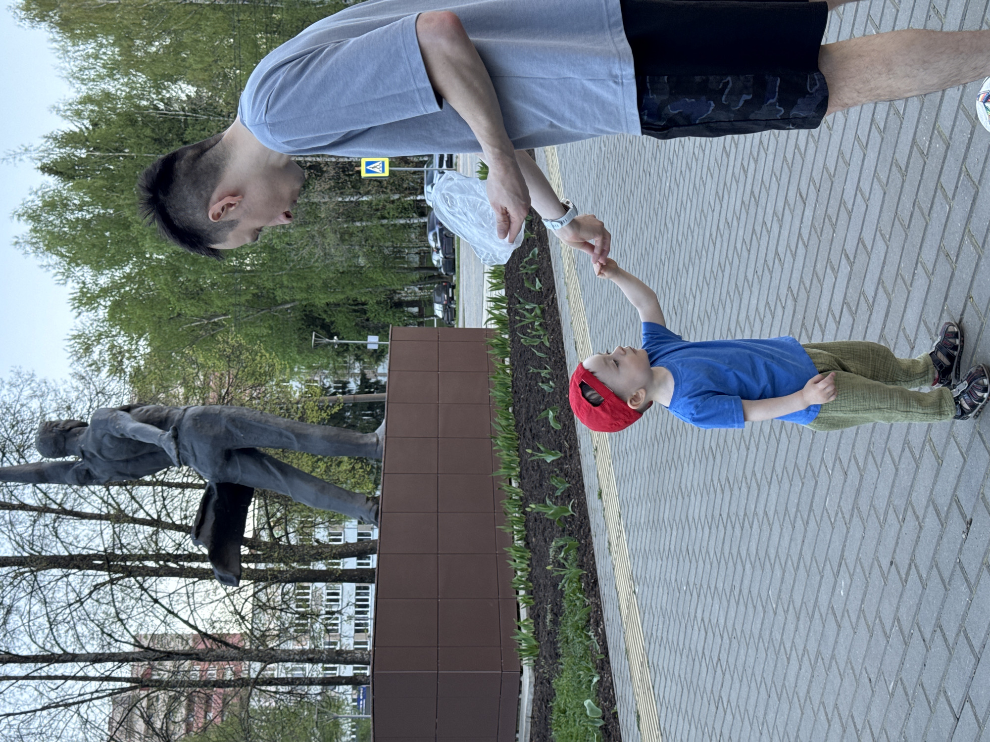 A man and a young boy are interacting on a brick paved area in Izhevsk, Russia. The man is wearing a gray t-shirt and black shorts with a pattern. He is holding a plastic bag in one hand and reaching down to touch the boy's hand. The boy, who is wearing a blue shirt, green pants, and a red cap, is reaching up towards the man. Behind them is a bronze statue of a man in a flying pose, mounted on a brown rectangular base. There is a row of flowerbeds in front of the base. Trees are visible in the background, along with buildings and a pedestrian crossing sign. The sky is overcast.