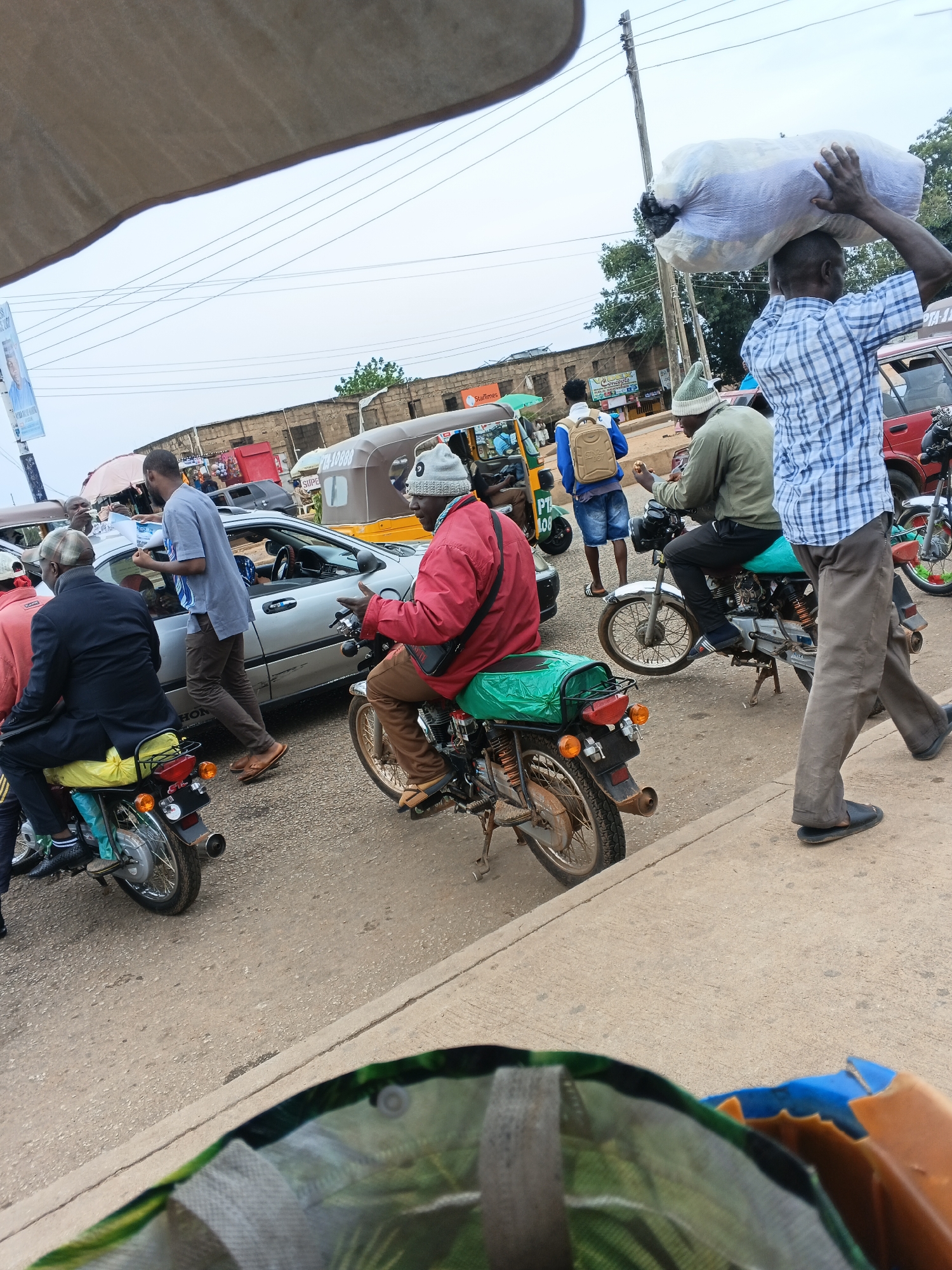 In Bukuru, Nigeria, a busy street scene unfolds with several individuals and vehicles. A man in a blue and white checkered shirt carries a large white bag of goods on his head, walking on the right side of the frame. He is dressed in brown trousers and dark sandals. To his left, a man wearing a red jacket and a grey beanie rides a motorcycle, also in brown trousers and sandals. His motorcycle has a green seat cover. Another man in a grey shirt and brown trousers stands next to a silver car, appearing to interact with someone inside the vehicle. In the background, a three-wheeled vehicle, possibly a tuk-tuk, is visible, along with other motorcycles and cars. The environment appears to be a commercial area with buildings and utility poles lining the street under an overcast sky.