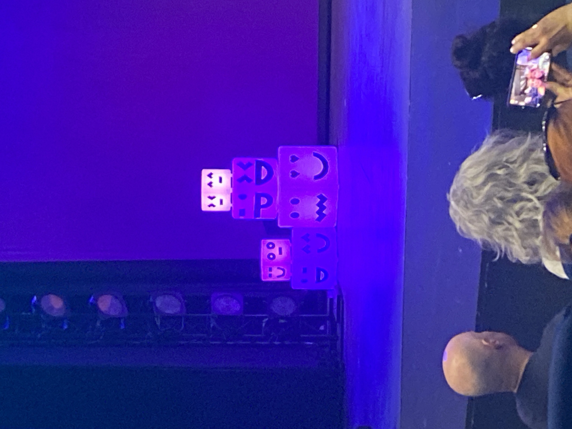 The image shows a stage with several lit cubes stacked on top of each other. The cubes have cartoonish faces lit from within, giving them a glowing effect. The stage is illuminated with purple and blue light. In the foreground, the backs of several audience members are visible. One person in the foreground is holding up a smartphone, appearing to take a photo or video of the stage. The overall atmosphere suggests a performance or event. The scene is not identifiable as being in Barcelona, Spain.