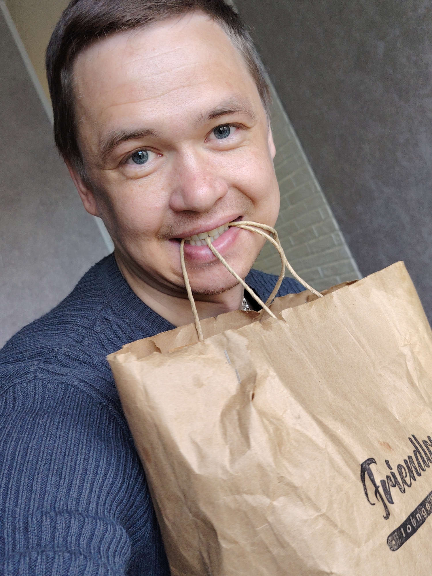 A man with light skin and blue eyes is holding a brown paper bag in front of him. He is biting the bag's handles with his teeth and smiling. He is wearing a dark blue sweater. The bag has the words "Friendly Lounge" printed on it in black ink. The background is a gray wall. The location is Chaikovsky, Russia.