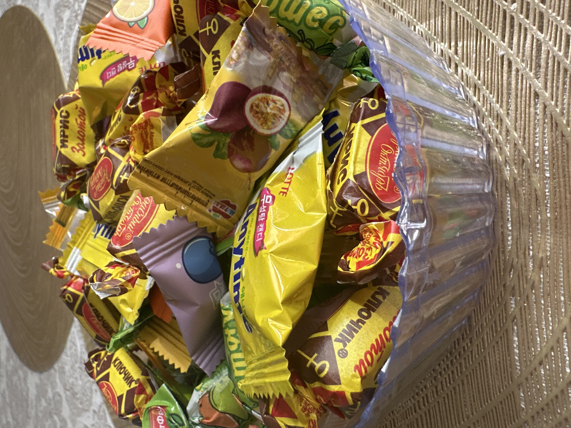 A close-up, eye-level, indoor shot shows a clear plastic bowl filled with an assortment of individually wrapped candies. The candies are predominantly yellow, orange, and purple, with some featuring red and brown accents. Many wrappers have visible text in Cyrillic and other scripts, as well as images of fruit, such as passion fruit and citrus. The candies are piled high, obscuring the bottom of the bowl. The bowl rests on a textured, beige placemat, which is placed on a light-colored surface, possibly a table. The background is a soft-focus, slightly blurred depiction of a light brown, patterned surface. The lighting suggests a warm, indoor environment, casting subtle shadows on the candies and the background. The scene is static, with no visible action or people. The specific location in the city of Khabarovsk, Russia cannot be determined from the image alone.