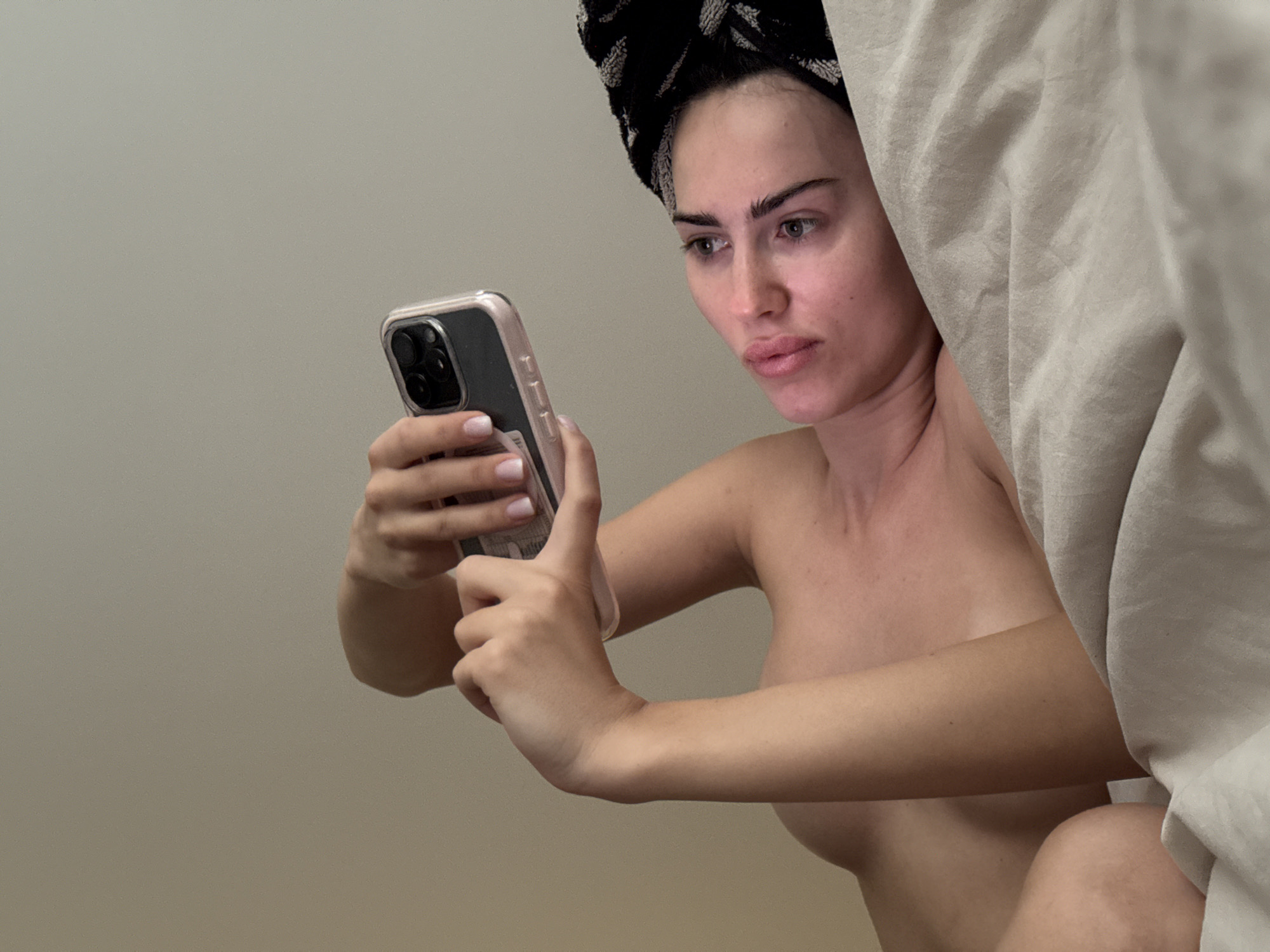 A woman with a towel wrapped around her head is holding a smartphone in her hands and looking at the screen. The woman is positioned on the right side of the image, near a white curtain. A plain wall is visible in the background on the left.