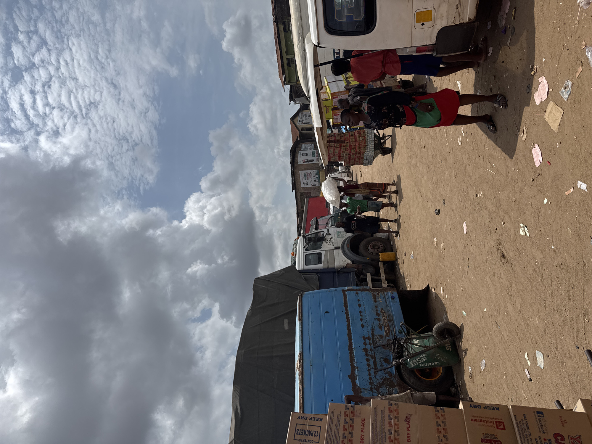 The image captures a street scene in Onitsha, Nigeria. The ground is sandy, littered with debris. In the foreground, there are stacks of cardboard boxes with "KEEP DRY" and "DRY PLACE" printed on them. Next to the boxes is a blue, rusted vehicle and a green wheelbarrow. Further into the scene, there are people walking and interacting near a white truck. Several buildings are visible in the background. The sky is filled with heavy clouds.