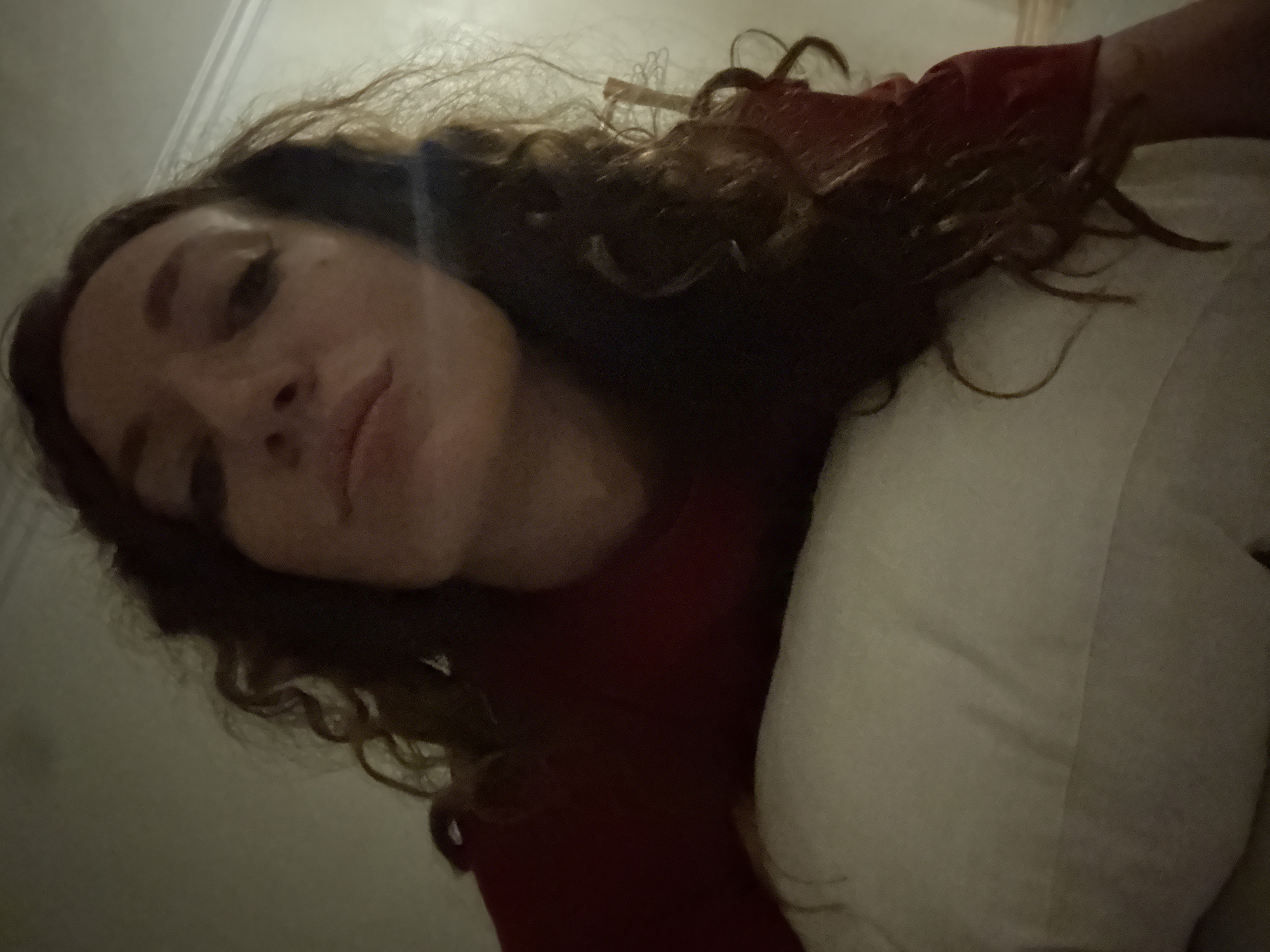 A low-angle, close-up shot captures a woman with long, curly brown hair wearing a red garment. She is lying on a white pillow, her head tilted back. Her facial features are visible, including her eyes, nose, and lips, with some makeup applied to her eyes. The background appears to be an indoor setting with a light-colored wall and a hint of molding at the top. There are no discernable landmarks or clues to indicate the location within Mai Chau, Vietnam.