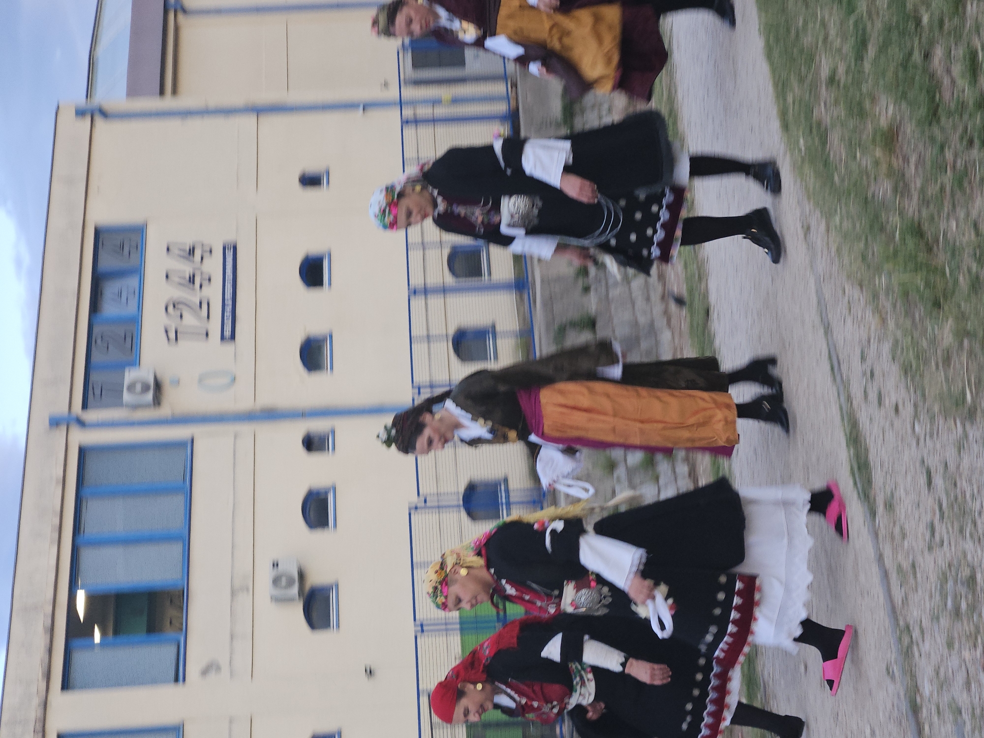 The image shows a line of six women in traditional dress, walking along a path. They are in front of a building with the number 1244 on it. The location is the city of Unknown, Unknown. The building is a light tan color with blue trim around the windows. The women are wearing a variety of colorful dresses. The path is made of concrete and there is grass on either side.