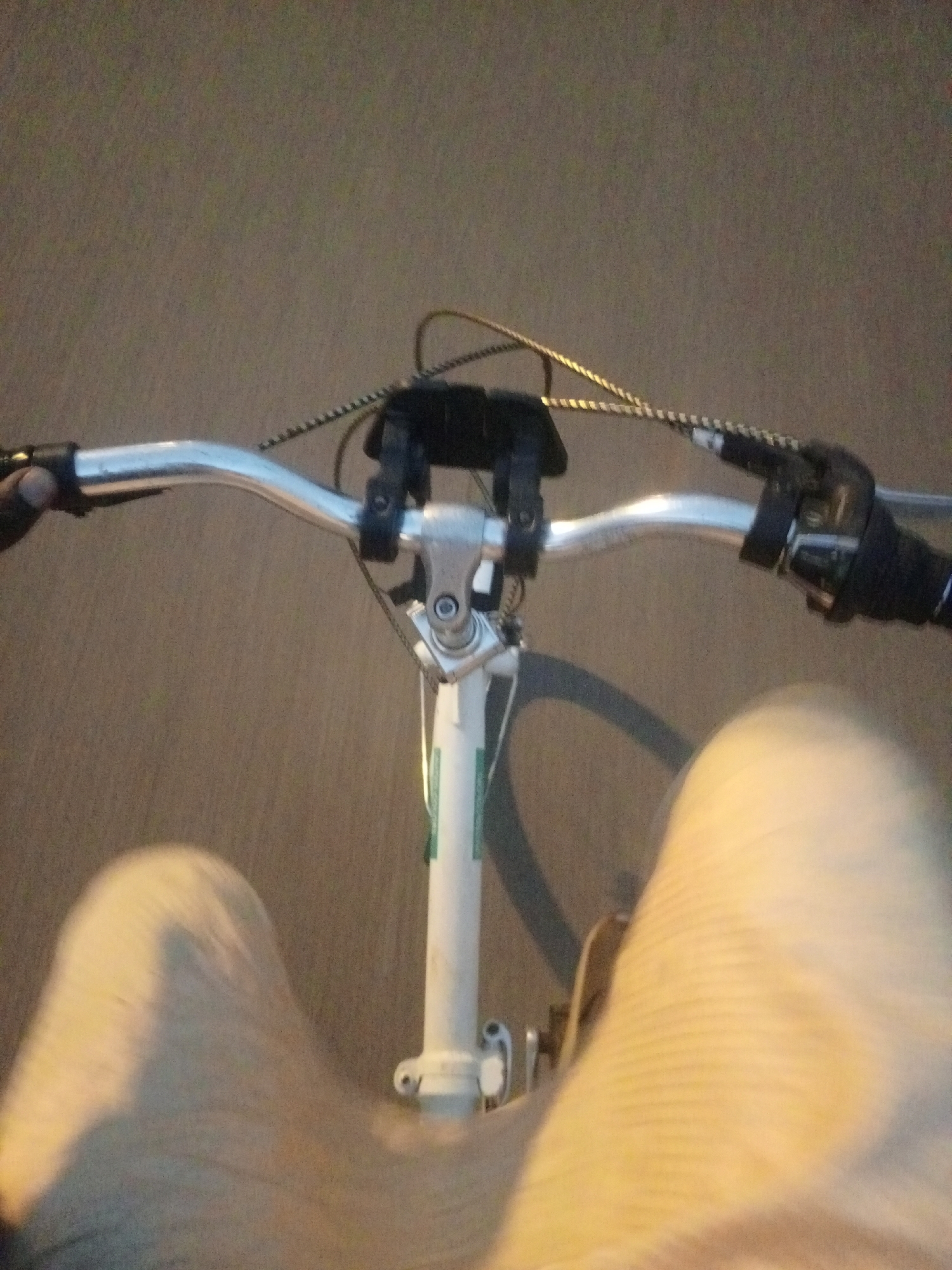 The image shows a first-person perspective of someone riding a bicycle. The handlebars, stem, and front fork of the bicycle are visible. The rider's legs, clad in light-colored pants, are also in the frame, with the left leg more prominent and in focus. The background is a blurred asphalt surface, suggesting the bicycle is being ridden on a road. The lighting suggests it might be dusk or early morning, or an area with artificial lighting. The image's composition and blur indicate movement. While the image itself does not provide any explicit indicators of location, the provided context states the location is Bukuru, Nigeria.