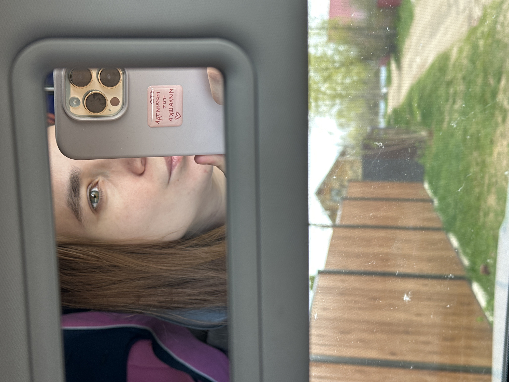 The image appears to be a selfie taken in a car, possibly in Chaikovsky, Russia (implied but not definitively proven). A person is visible reflected in a car mirror, holding an iPhone with a pink case. A sticker on the case has lettering in a non-Latin script. Outside the car window, a blurred scene includes a wooden walkway or steps, greenery, and a building. The image is somewhat distorted by the angle and reflection.