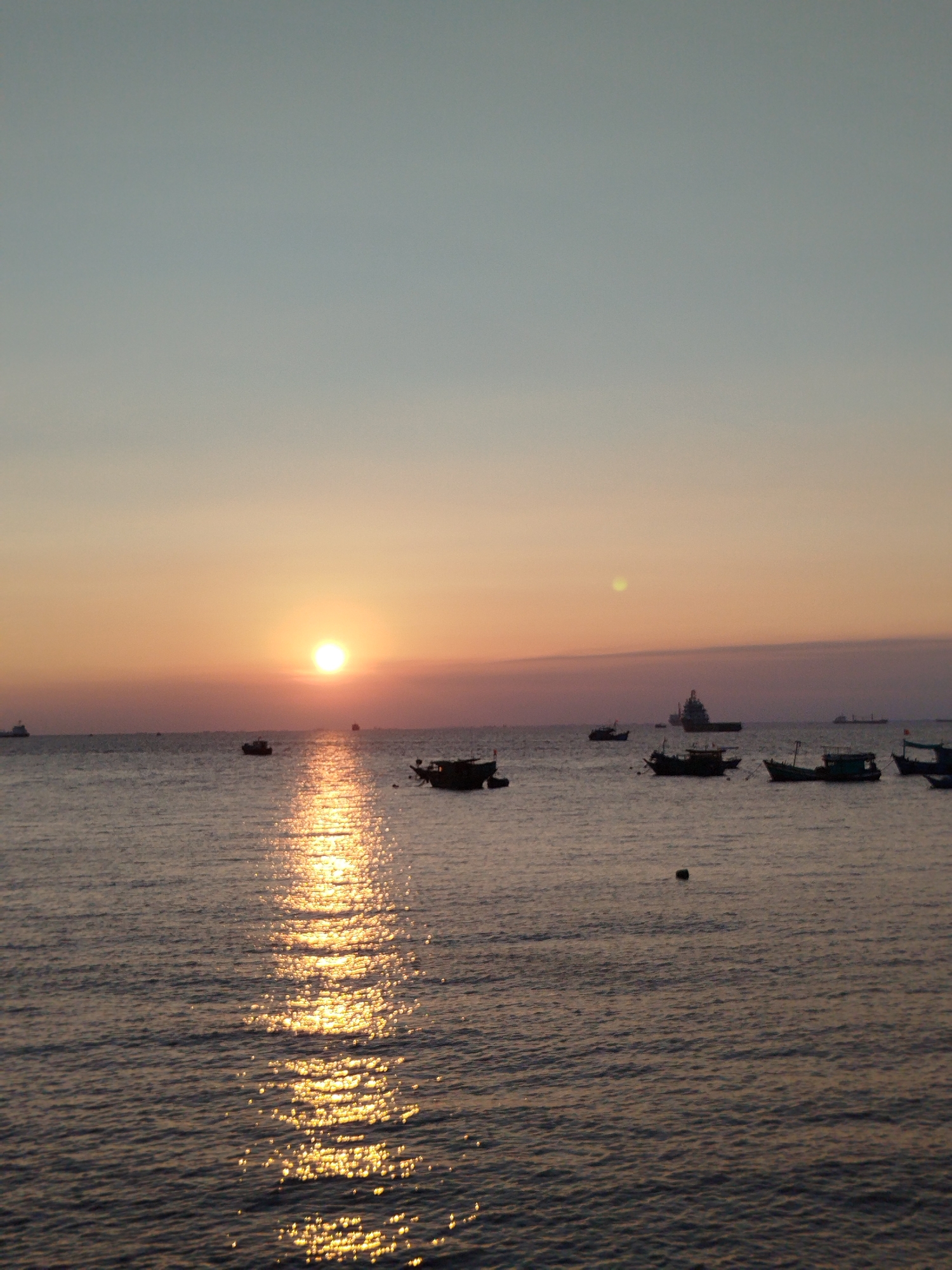 The image depicts a sunset over the sea, with several boats visible in the water. The sun is low on the horizon, casting a golden glow across the sky and reflecting on the water's surface. The location is likely coastal, near Ho Chi Minh City, Vietnam, based on the typical coastal scenery in that area. There are no people visible in the image.