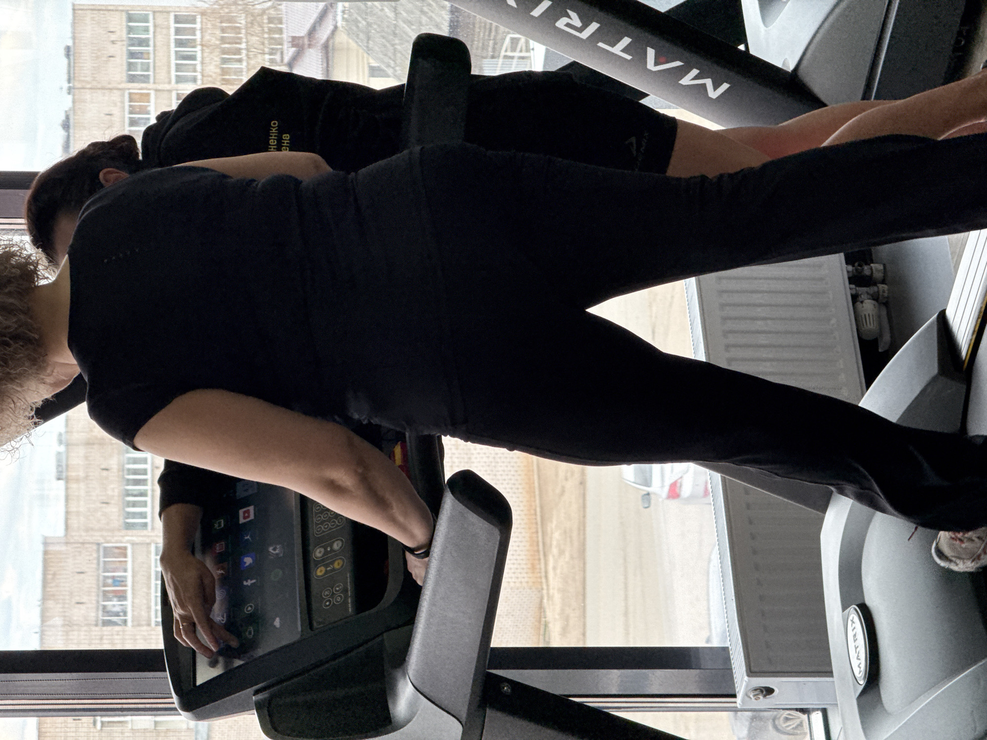This media file presents an indoor scene, likely a gym or fitness center, captured with a 90-degree clockwise rotation. Two individuals are depicted using treadmills against a backdrop of a large window overlooking an urban environment.
In the immediate foreground, a person with curly, light-colored hair is operating the console of a "MATRIX" brand treadmill. They wear a black short-sleeved top, and their right hand is shown touching the digital screen, which displays various application icons such as YouTube, Facebook, and Puma.
Behind this individual, a second person is visible from the back, dressed in black athletic wear, including what appears to be a long-sleeved top or hoodie and pants. Their dark hair is tied up. Partially visible Cyrillic text is embroidered on their back, appearing as "НЕНКО" on an upper line and "ОНЯ" on a lower line. This person is also on a "MATRIX" treadmill, with their right foot, clad in a white sneaker, visible on the running belt.
Through the expansive window, the exterior view reveals several brick buildings with multiple windows, suggesting apartment complexes or offices. A white vehicle can be seen on a road or parking area outside. The lighting indicates daytime, likely overcast, as no distinct shadows are cast. A white radiator is visible indoors beneath the window. The scene captures individuals engaged in physical activity on exercise equipment, in Votkinsk, Russia.
