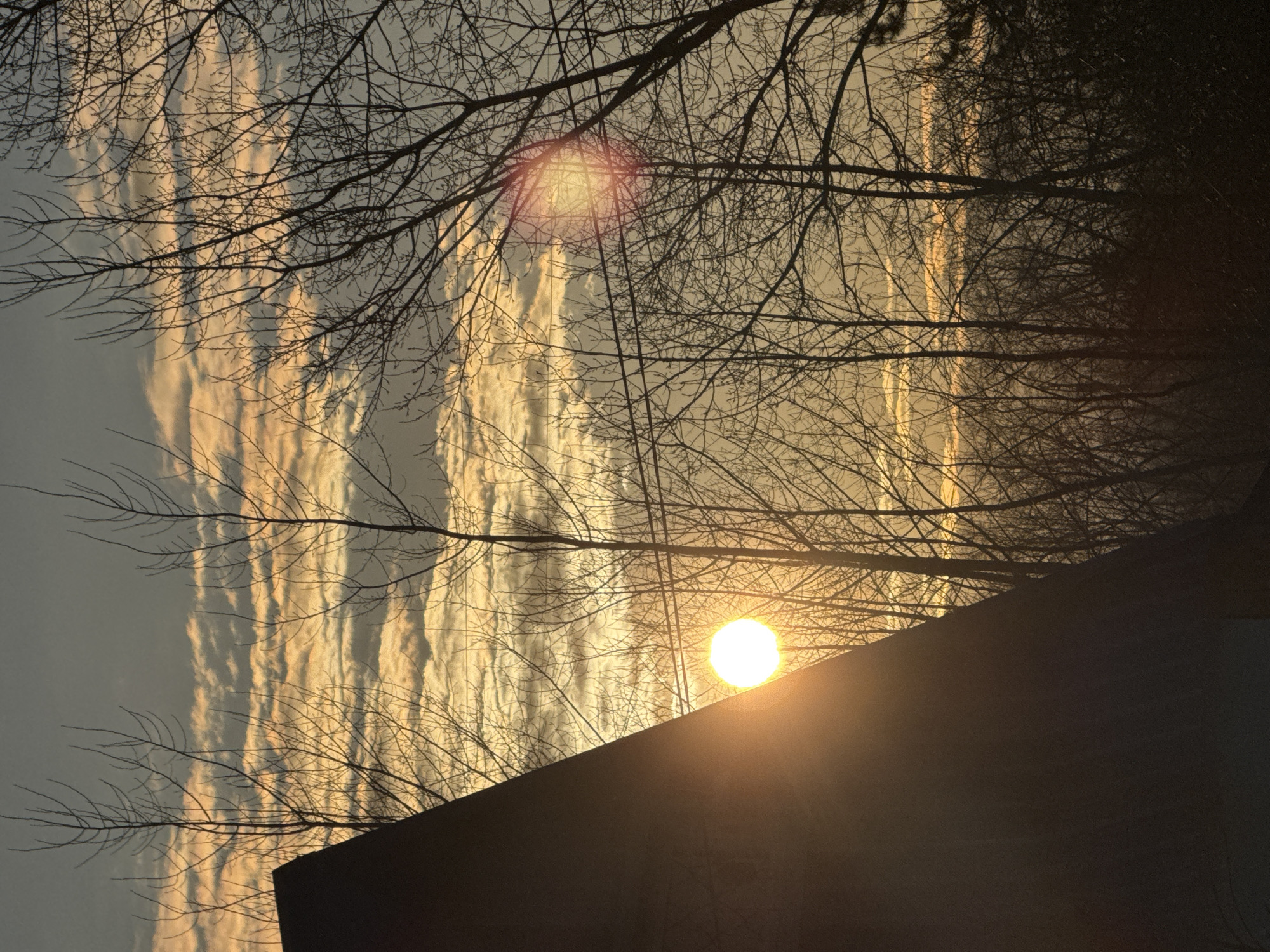 The image depicts a sunset in Chaikovsky, Russia. Silhouetted tree branches are visible against the sky with long vertical clouds illuminated by the setting sun. The bright sun is partially obscured by the silhouetted roofline of a building.