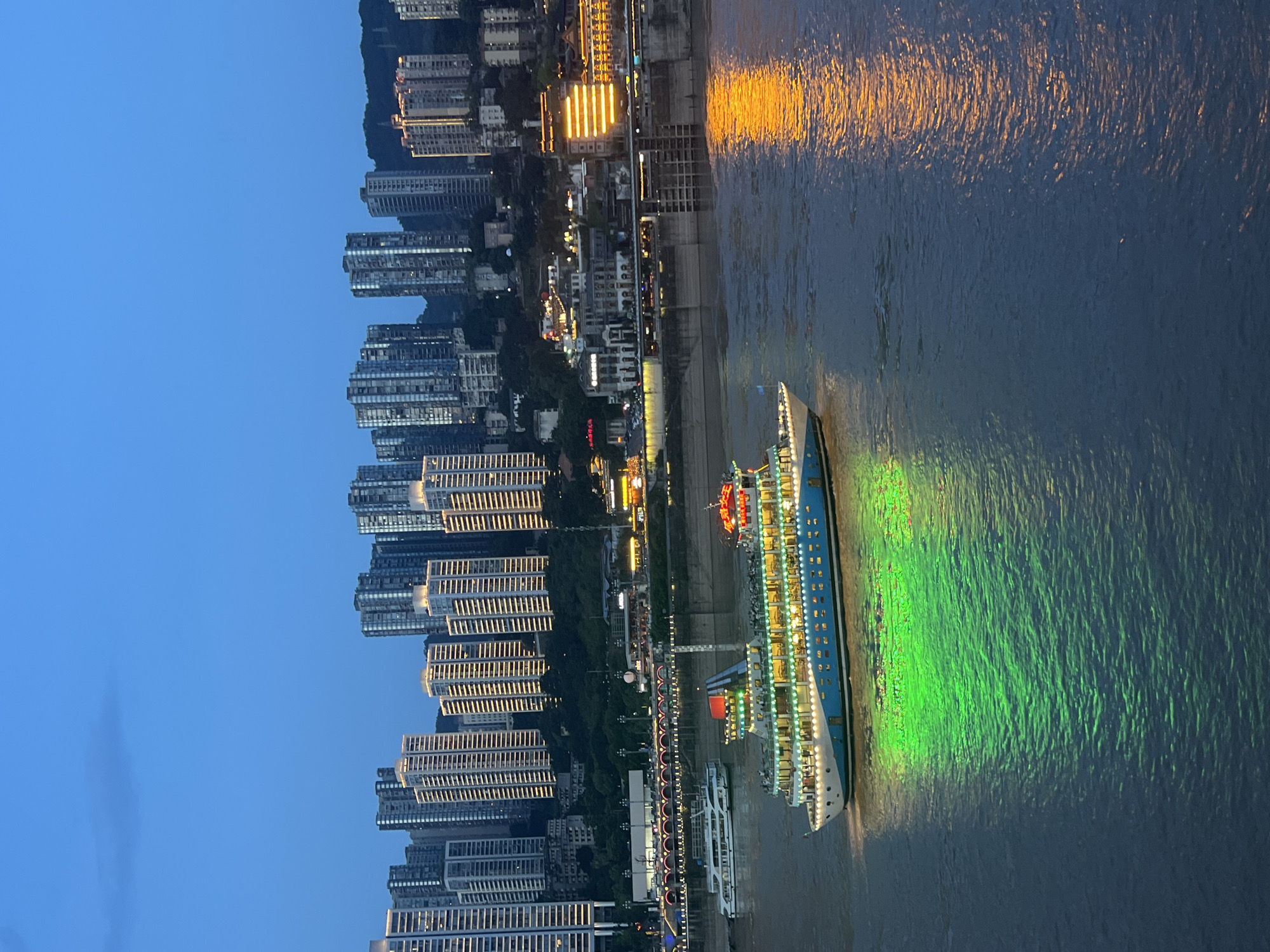 The image captures a twilight scene on the Yangtze River in the Yuzhong District of Chongqing, China. A brightly lit cruise ship, adorned with green and blue lights, sails on the water, casting a vibrant green reflection. In the background, the cityscape of Yuzhong District is illuminated with numerous buildings and structures, their lights twinkling against the darkening sky. The river is calm, reflecting the lights of the city and the ship. Another, smaller white boat is visible in the distance. The overall atmosphere is one of urban beauty and vibrant nightlife.