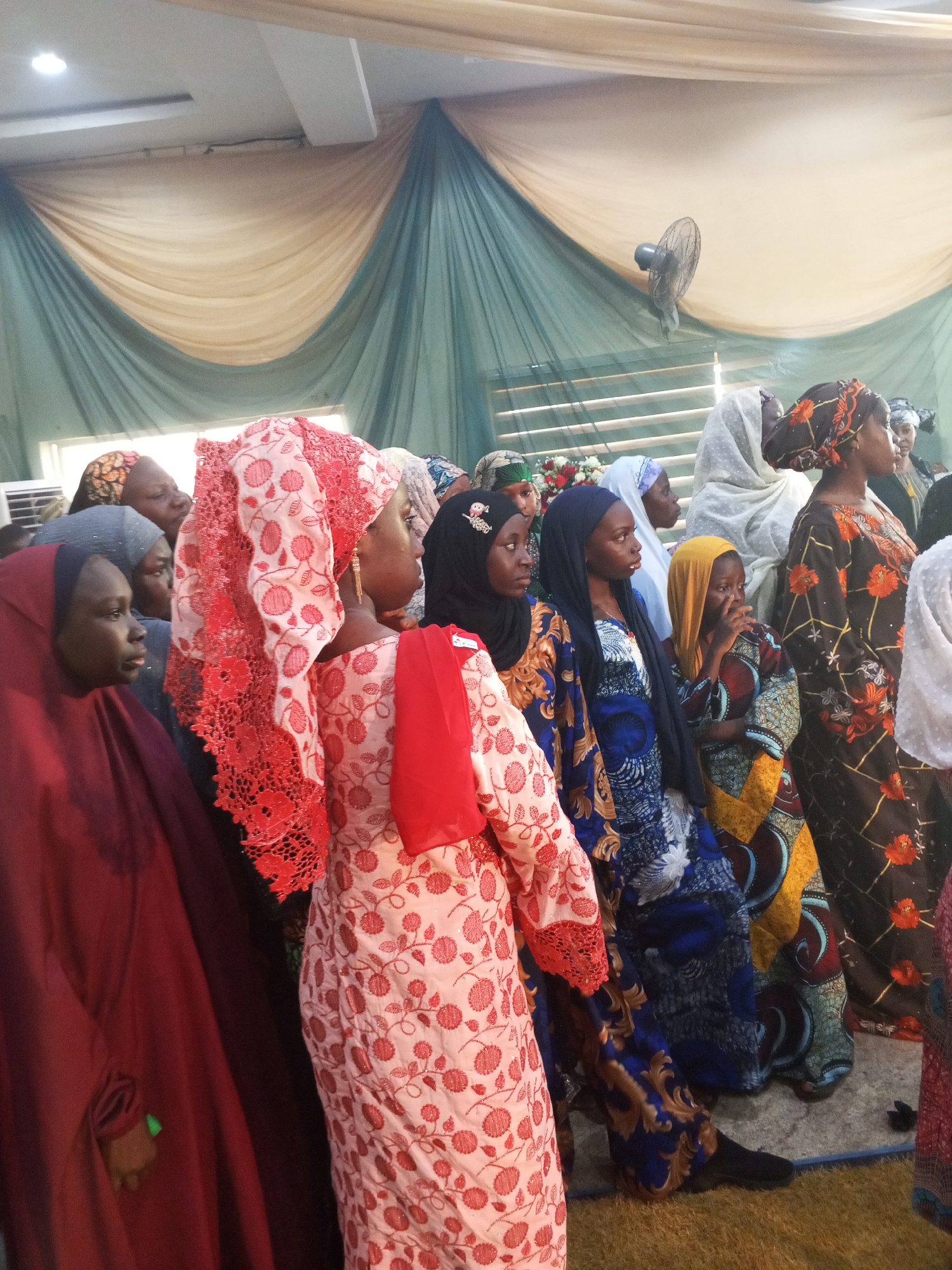A group of women wearing traditional African attire and hijabs are standing in an indoor setting. The women are mostly facing to the right of the frame, with some looking slightly off-camera. The clothing features vibrant patterns and colors, including red, pink, blue, yellow, and black. One woman in the foreground on the left wears a dark red hijab and abaya. Next to her, a woman in a pink abaya with red floral patterns and a red lace headscarf is prominent. Other women are dressed in patterned dresses and head coverings. The background consists of draped fabric in beige and green. A ceiling fan is visible mounted on the wall in the upper right portion of the image. The floor appears to be covered with a rug and some grass-like material. The lighting suggests an indoor event or gathering.