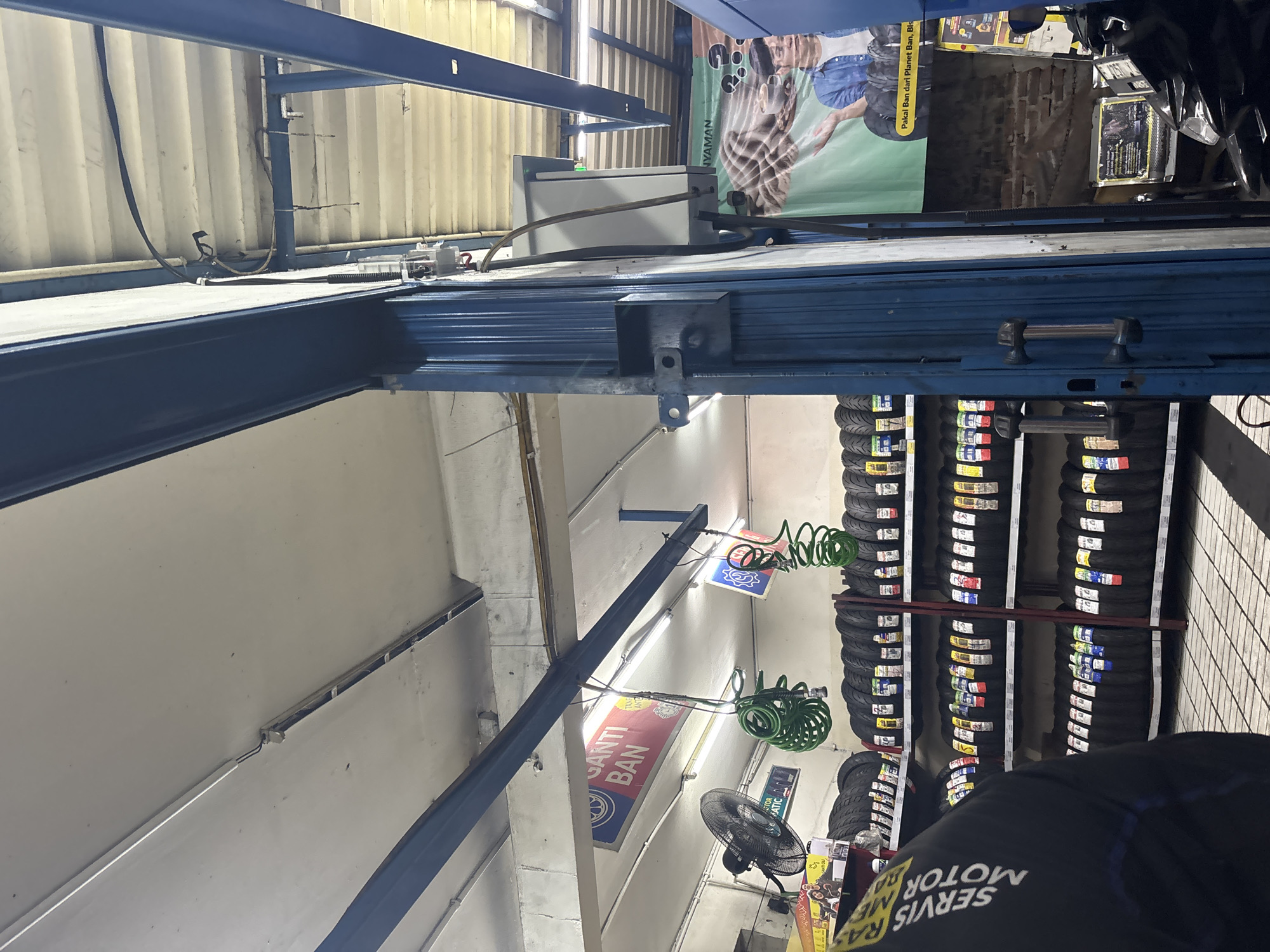 The image depicts the interior of a tire shop in North Jakarta, Indonesia, likely located on a street-level storefront. The scene is cluttered with stacks of motorcycle tires arranged on metal shelves that line the walls. Above, fluorescent lights illuminate the space, with some wires and hoses visible. A blue metal frame structure runs through the center of the image, supporting some of the ceiling elements and electrical conduits.
In the foreground, a person wearing a dark jacket with white lettering that reads "SERVIS MOTOR" is partially visible, suggesting they are an employee. To the left, a black oscillating fan is mounted on a shelf. Hanging from the ceiling are green coiled hoses, likely for air compressors.
Several signs are displayed: a red and blue sign with "GANTI BAN" (Change Tire) is prominent. Another banner in the background features a man and text that reads "Pakai Ban dari Planet Ban, Bls".
The overall environment suggests a functional workshop focused on selling and servicing motorcycle tires. The lighting is a mix of artificial light from the overhead fixtures. The floor appears to be tiled.