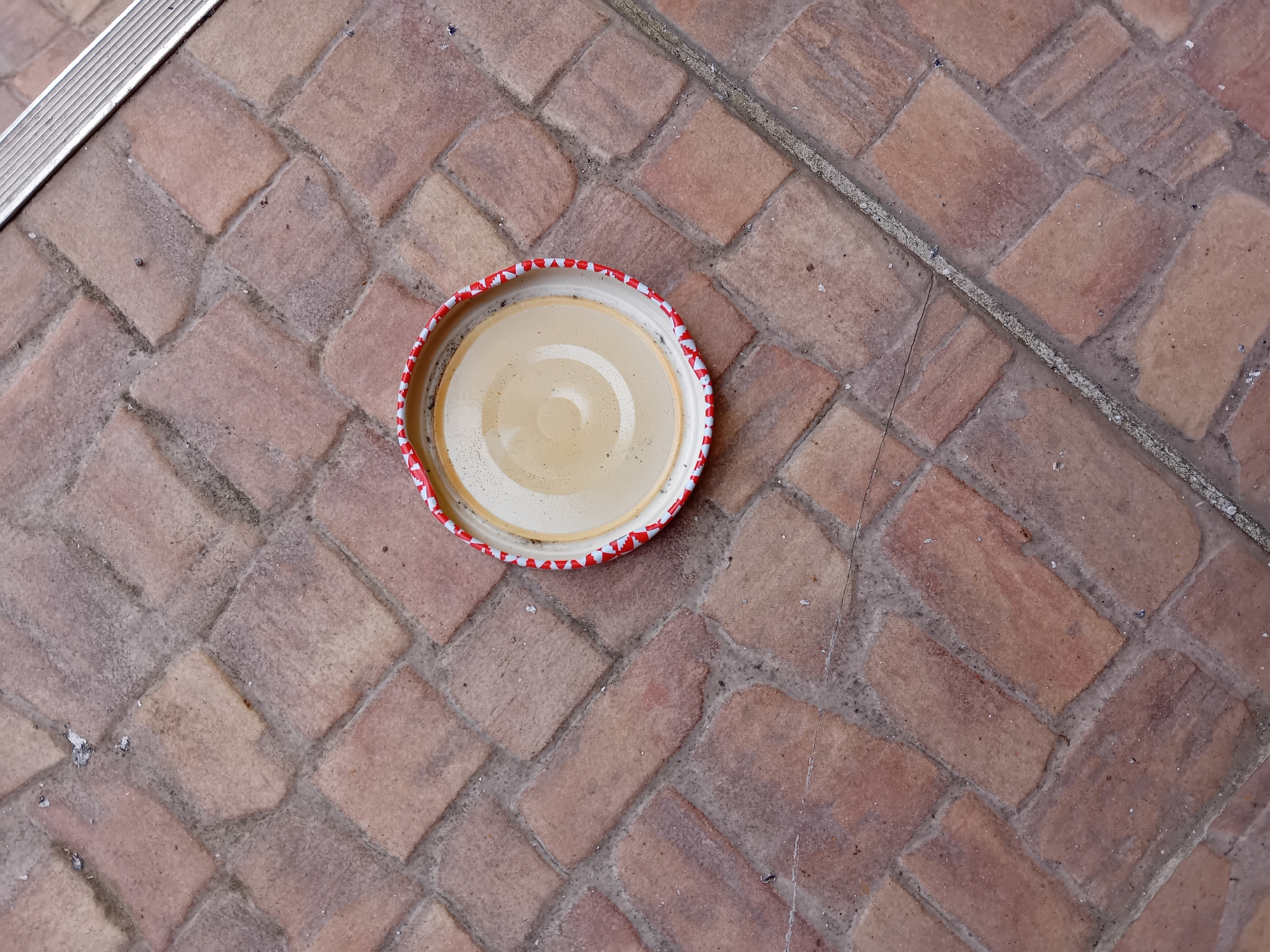 An overhead shot shows a red and white patterned bottle cap resting on a tan brick patio. The cap is circular and faces upward, revealing a light tan interior. The brick patio is laid in a herringbone pattern, with a metal strip running along the top left edge of the image and a grout line diagonally across the upper right portion of the frame. The bricks are rectangular and a reddish-brown in color. The scene appears to be outdoors in a common area of Zuberec, Slovakia. There are no people or animals visible in the image, and no discernible actions or interactions are taking place. The lighting is natural and casts faint shadows.