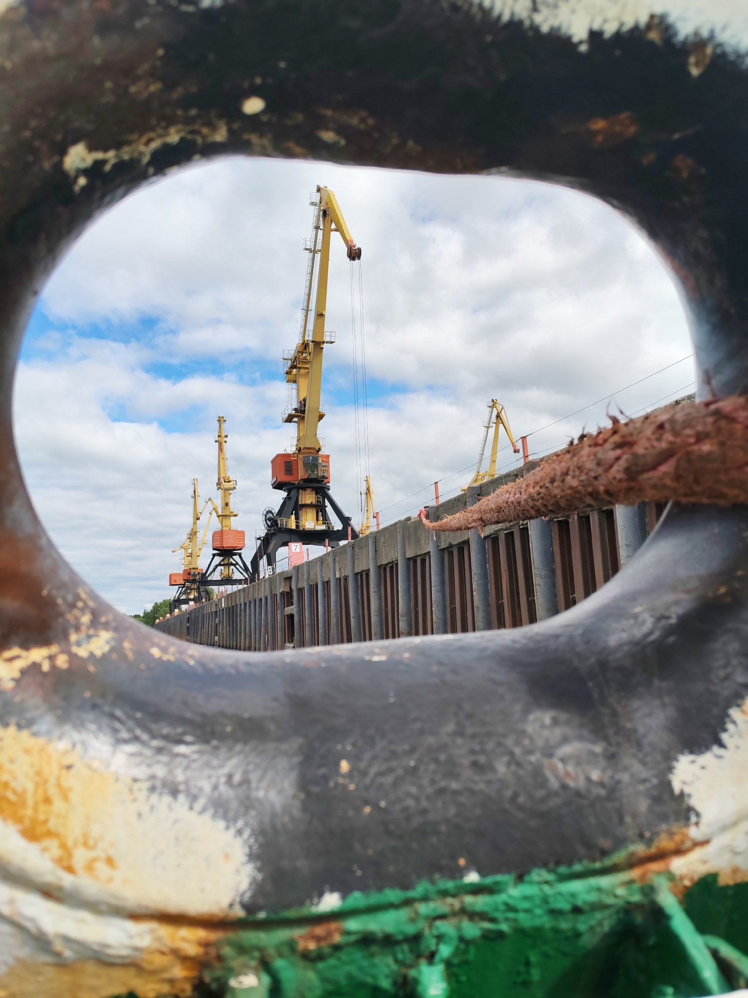 The image shows a view of cranes at the port of Rybinsk, Russia, as seen through a mooring ring. The ring is large, painted black with rust and flaking paint showing yellow, green, and white underneath. A rope is tied to the ring. The cranes are yellow and black, with orange accents. They stand behind a concrete and metal barrier at the edge of the dock. The sky is cloudy with patches of blue.