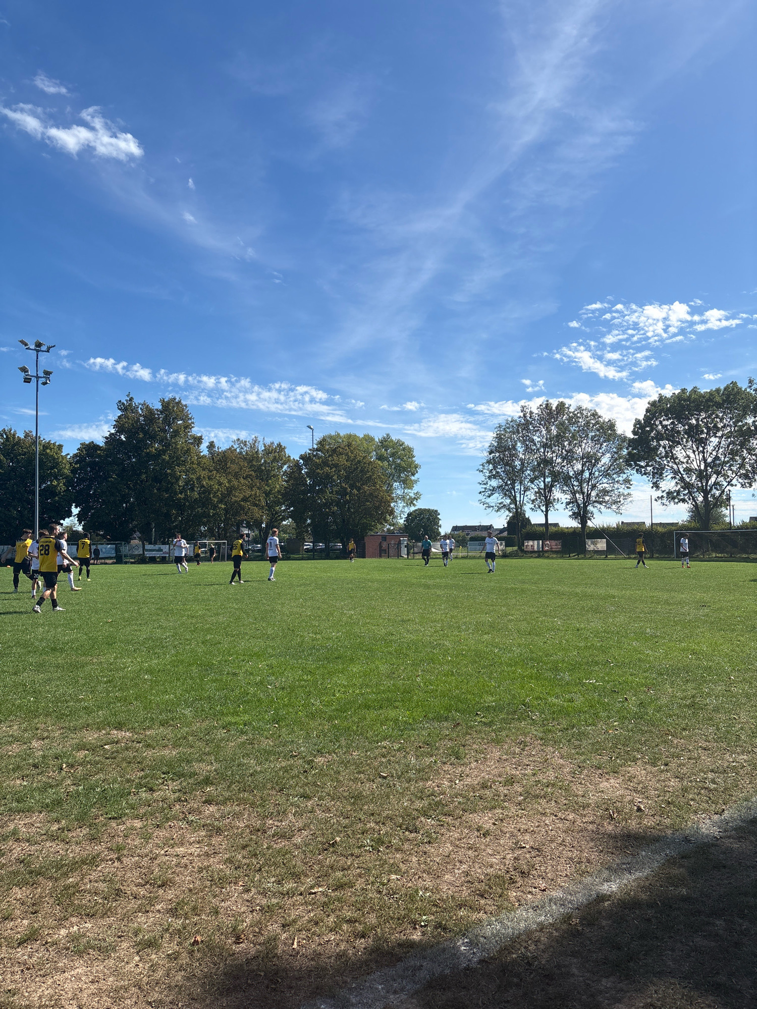 The image depicts a soccer match taking place on an outdoor field. Several individuals, presumably players, are scattered across the grassy field, some in motion, suggesting an active game. They are wearing athletic attire, including jerseys and shorts, in various colors, predominantly white and yellow, and black. The field itself exhibits patches of dry, brown grass mixed with greener areas, particularly towards the foreground. A soccer goal is visible on the right side of the frame. In the background, trees with green foliage form a natural boundary, and beyond them, a few buildings and streetlights can be observed. The sky above is a clear blue with scattered white clouds. Based on the provided information, this scene is located at the Sportanlage Am Bahndamm in Wegberg, Germany.