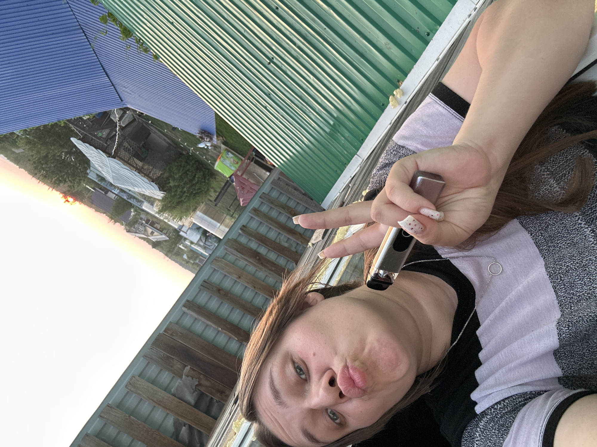 A selfie shows a person making a kissy face and holding a vaping device in their right hand, along with a peace sign. They are wearing a gray and black striped shirt and a silver necklace. The background reveals a residential area under construction in Ольховка, Russia. A green metal roof is visible in the upper right, and a blue metal roof is visible in the upper left. A wooden fence and a partially constructed building are in the mid-ground. In the distance, there are houses and trees silhouetted against a twilight sky.