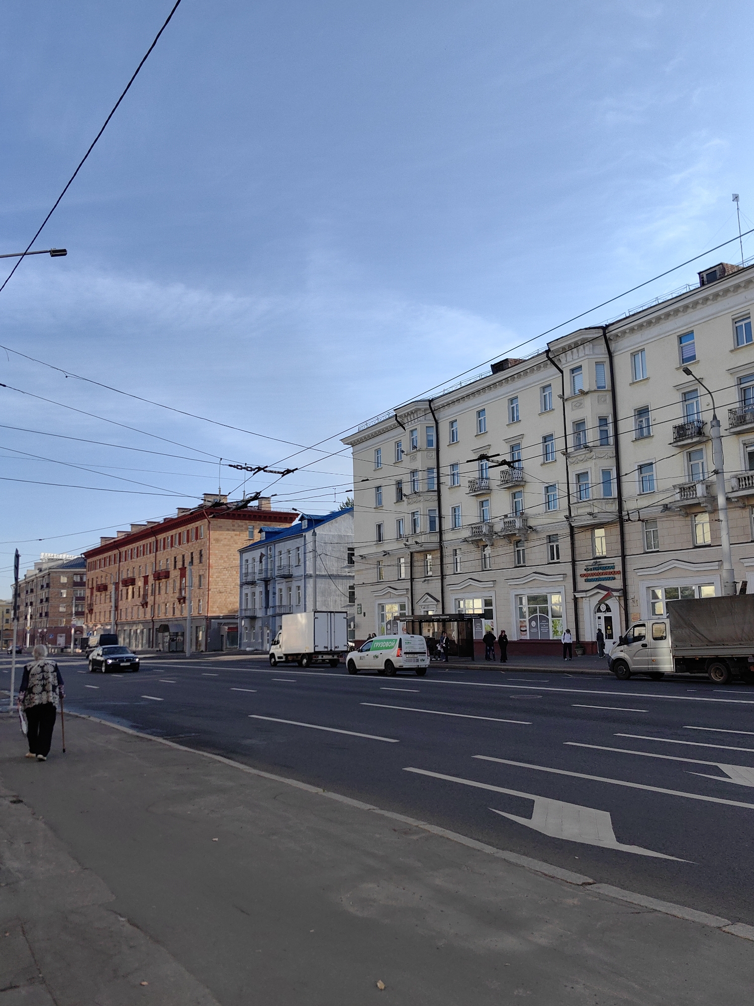 The image shows a street scene in Minsk, Belarus, possibly on Independence Avenue. The sky is a clear, pale blue with faint clouds. A woman with gray hair, wearing a dark patterned top and black pants, walks along the sidewalk with a cane. She carries a bag in her left hand. In the background, there are several multi-story buildings with light-colored facades and ornate architectural details. Some buildings have balconies and arched windows. There are overhead wires for what appears to be a trolleybus system. Cars are driving on the road, including a black sedan and a white truck. A white van with green lettering is parked on the right side of the road. Street markings on the asphalt indicate traffic lanes and directional arrows. A bus stop shelter is visible near one of the buildings. The scene depicts a typical urban environment with pedestrian and vehicular traffic.
