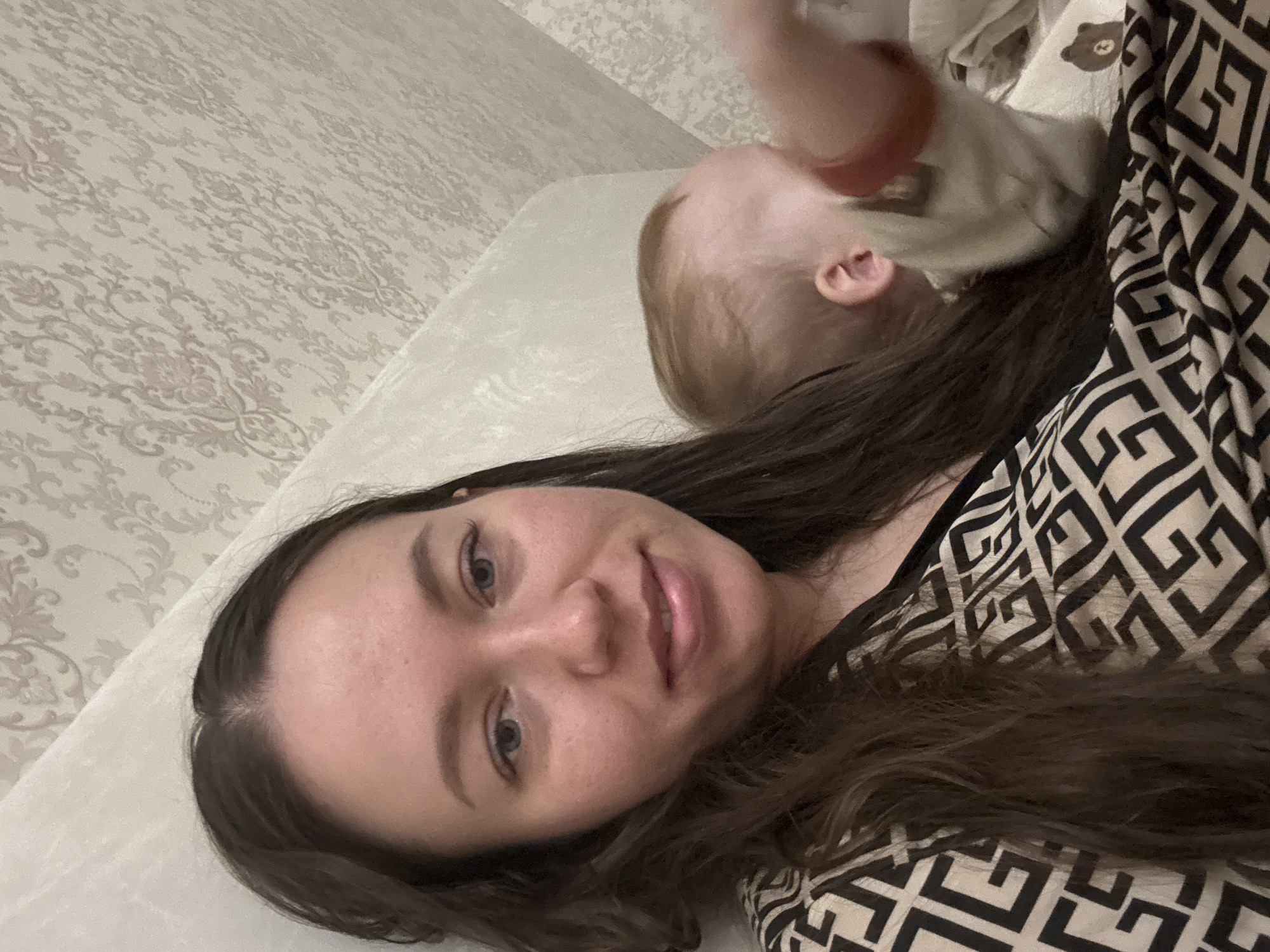 A woman with long brown hair is lying on a light-colored bed with a patterned wallpaper. A baby in a beige shirt is positioned near her head. The woman is wearing a patterned top. The image may have been taken indoors in the location of Ферма, Russia.