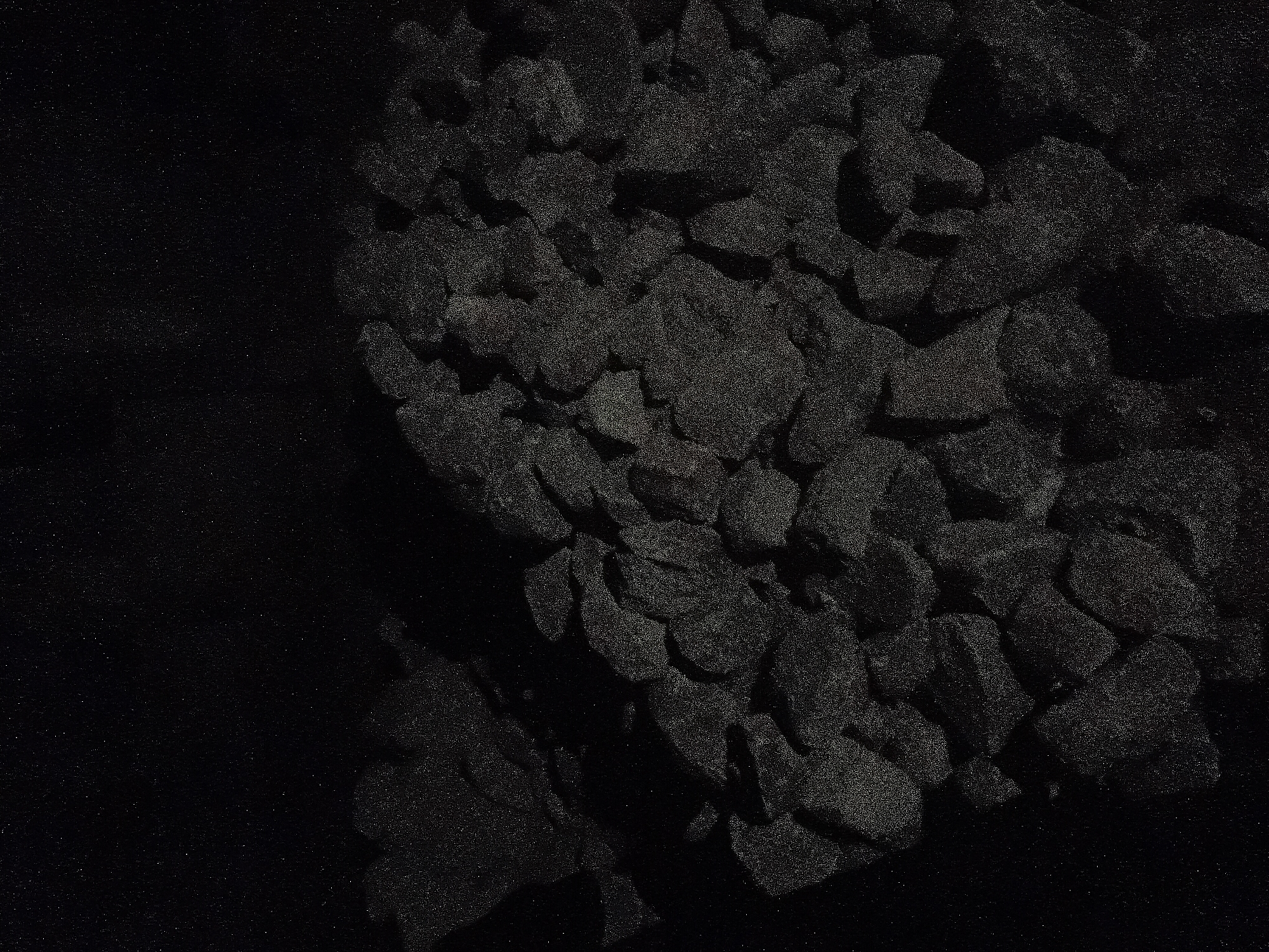 The image displays a dark, grainy, low-light scene featuring a large, irregularly piled mass of angular, dark grey to black granular material, consistent with crushed rock, gravel, or coal. The individual pieces are varied in size and shape, creating a rough, textured surface with subtle highlights and deep shadows indicative of low illumination. The material occupies the lower two-thirds of the frame, receding into a uniformly dark and featureless background in the upper portion. No human subjects, identifiable structures, or specific environmental markers are present to provide scale or further context. This scene is located in the city of Jalingo, Nigeria.