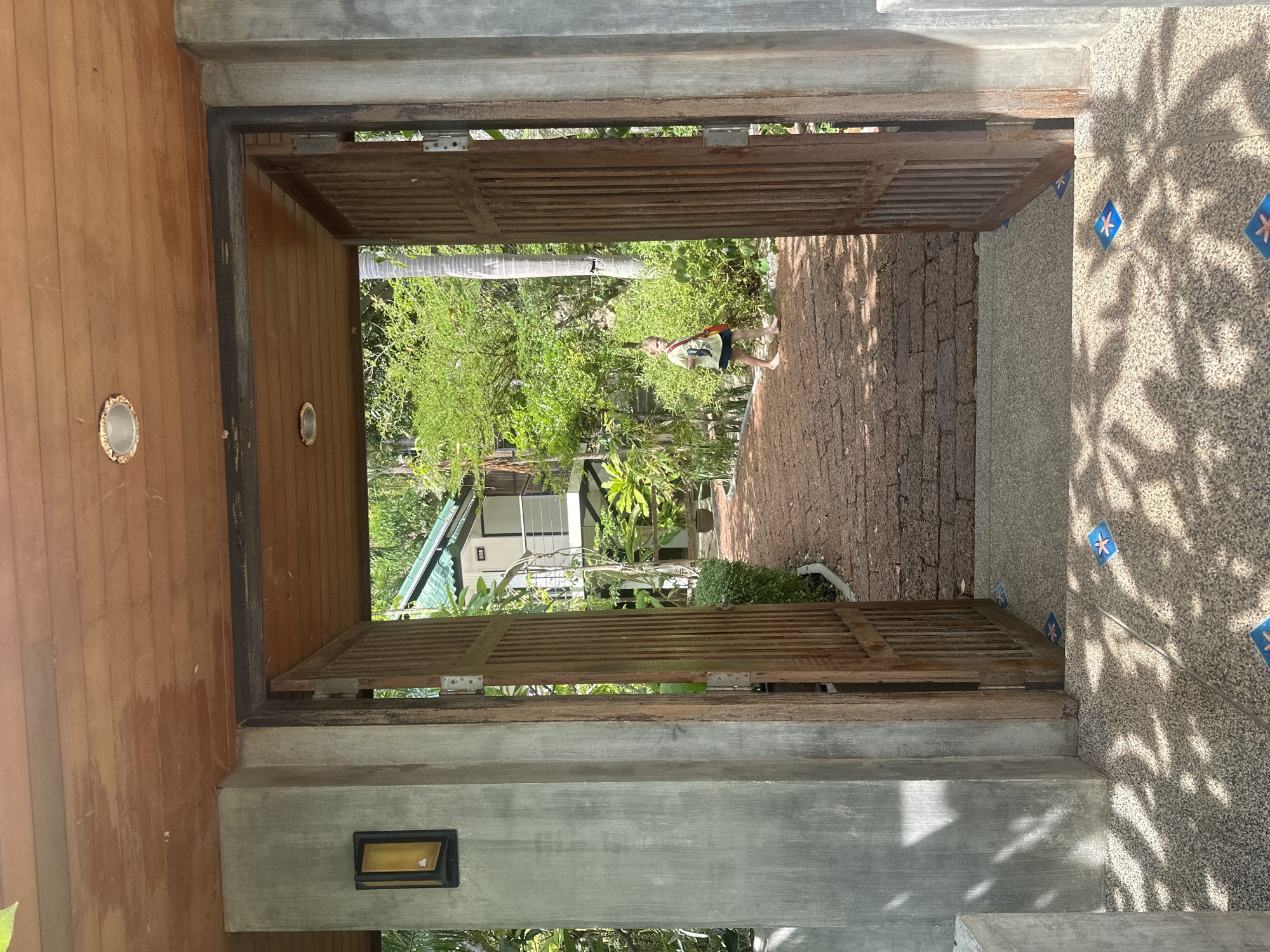 An open doorway reveals a young boy walking on a brick pathway in a tropical environment. The boy, wearing a yellow shirt and dark shorts, carries a red bag and walks barefoot. The pathway leads towards a white building with a green roof, surrounded by lush greenery and palm trees. The doorway itself is constructed from concrete and wood, with ornate wooden gates. The foreground shows a patterned tiled floor and dappled sunlight. The scene appears to be in a resort or private residence in ตำบลเกาะพะงัน, Thailand.