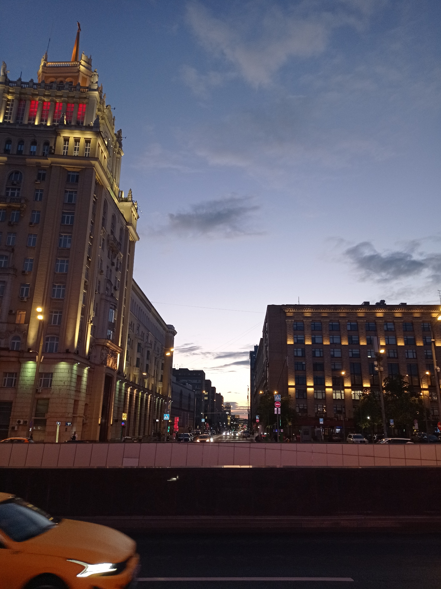 The image depicts a twilight view of a street in Moscow, Russia, likely in the evening. On the left, a prominent Stalinist Empire-style building dominates the scene, its upper floors illuminated with red lights, and its facade adorned with architectural details. Streetlights line the side of the building, casting a warm glow. Across the street, another building, also illuminated with warm lights, shows a grid of windows. Cars are visible on the street, suggesting ongoing traffic. In the foreground, a yellow car is partially visible, its headlights on, indicating motion. The sky is a dusky blue with scattered clouds, typical of the transition from day to night. The overall impression is an urban streetscape at dusk, characterized by imposing architecture and the ambient light of the city.
