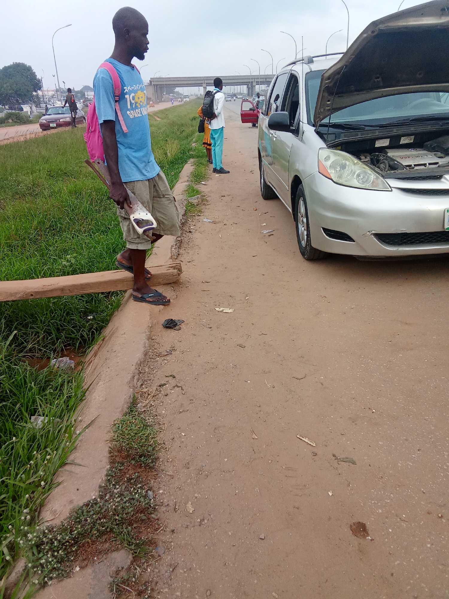 In Kagini, Nigeria, a man stands on a wooden plank crossing a shallow ditch near a road. He wears a blue t-shirt with a pink backpack and khaki shorts, and holds a saw. To his right, a silver minivan with its hood open is parked on the side of the road, suggesting a mechanical issue. The background shows other vehicles, a bridge, and vegetation, indicating an urban or semi-urban setting with ongoing traffic. The sky is overcast.